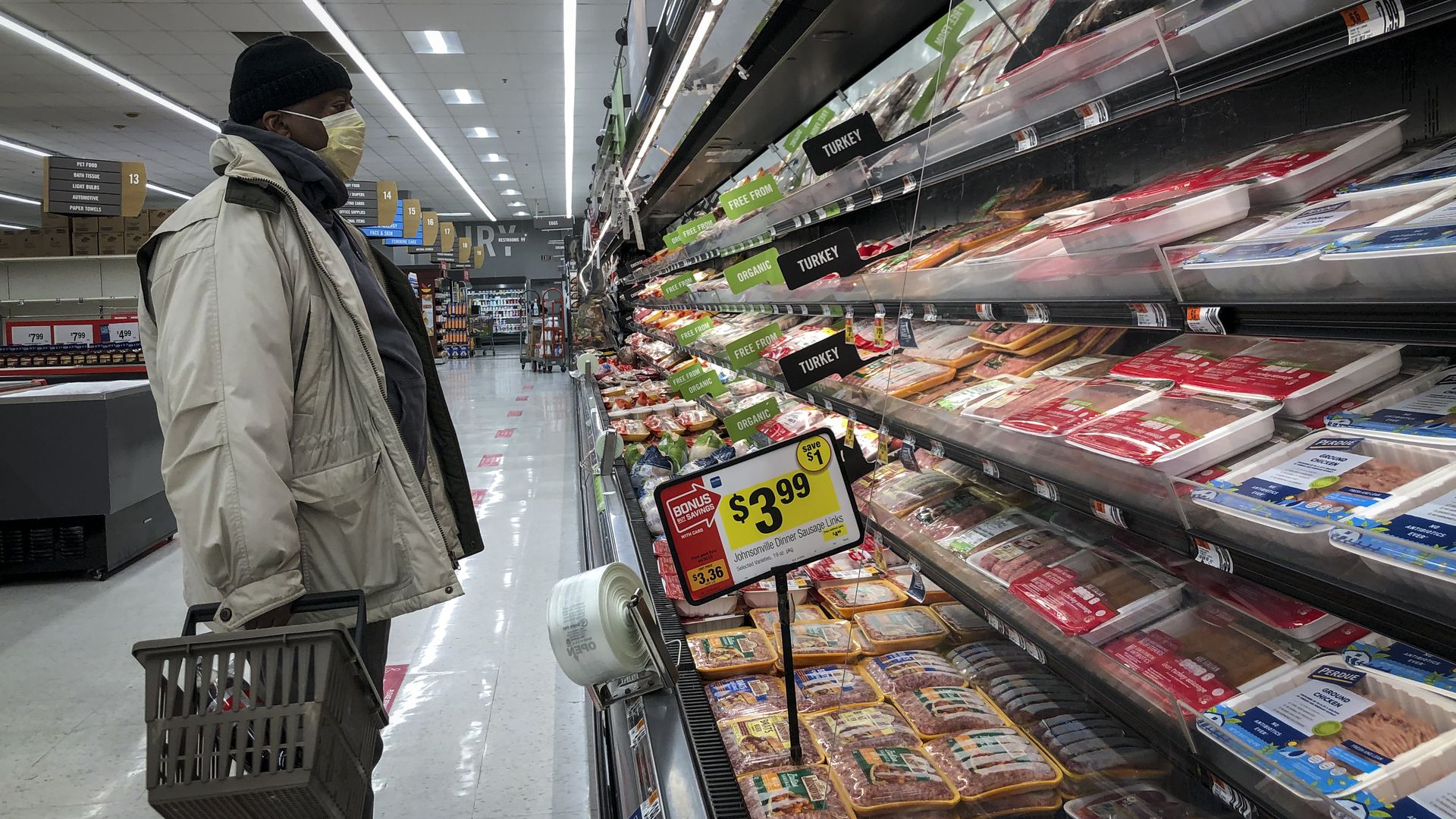 In this image, a man stands with a shopping basket in front of a meat produce aisle 