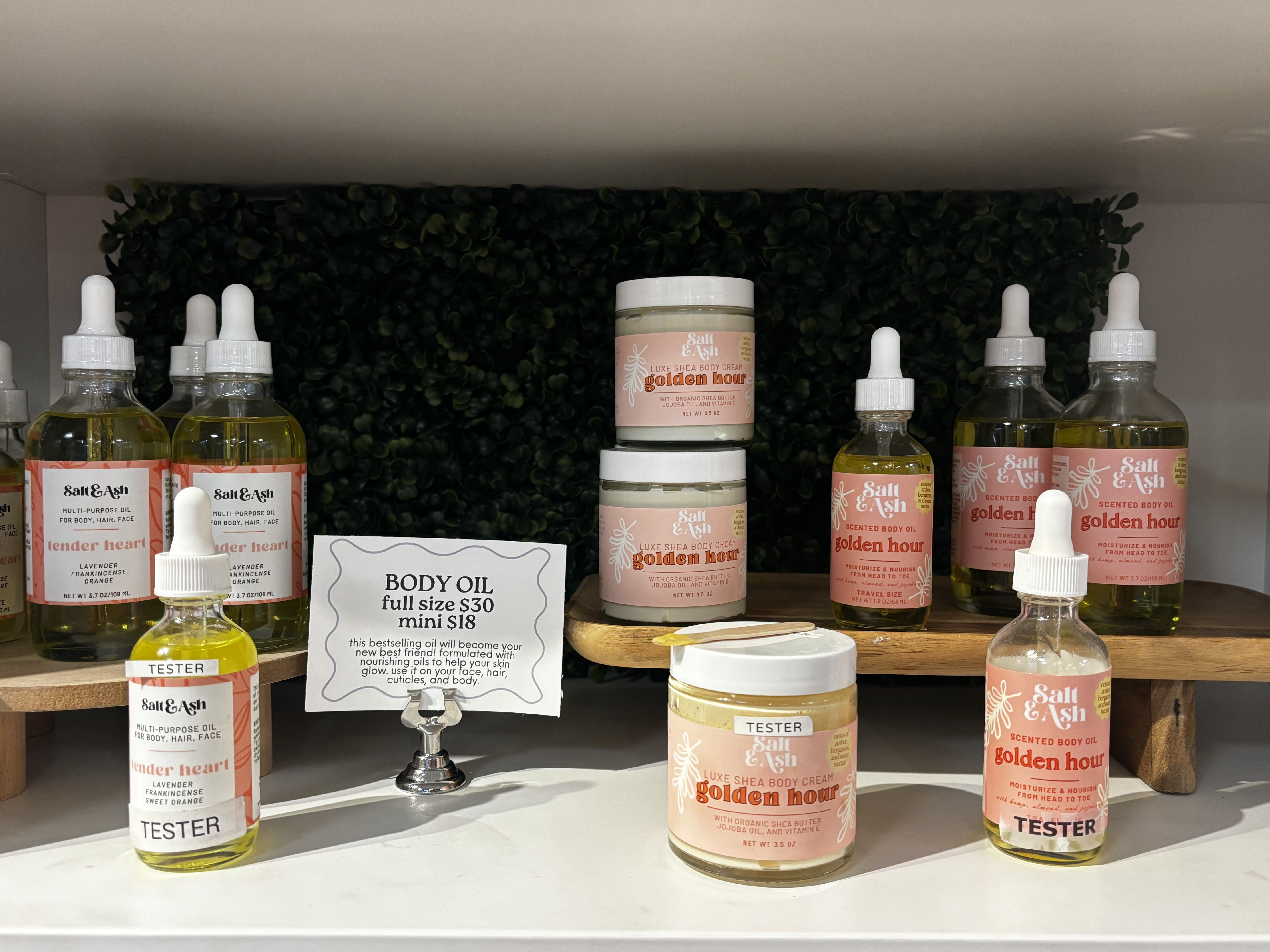 Display of Salt & Ash body oils and creams in pink-labeled bottles and jars, including "Golden Hour" and "Tender Heart" varieties, with a sign showing prices $30 full, $18 mini.