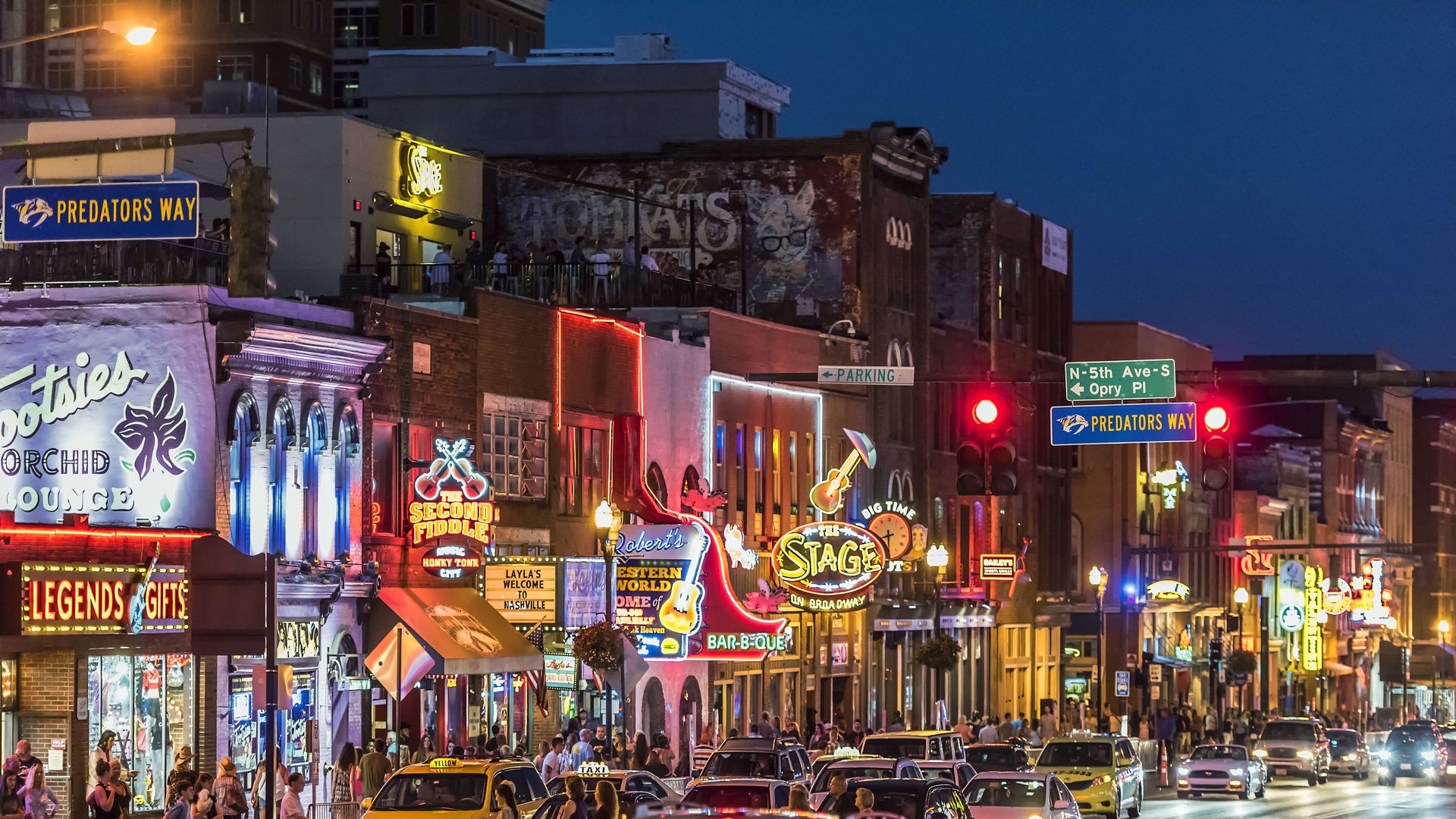 Nighttime neon-lit city street crowded with pedestrians, taxis, and colorful storefront signs on brick buildings; blue Predators Way signs and bright venue marquees line the block.