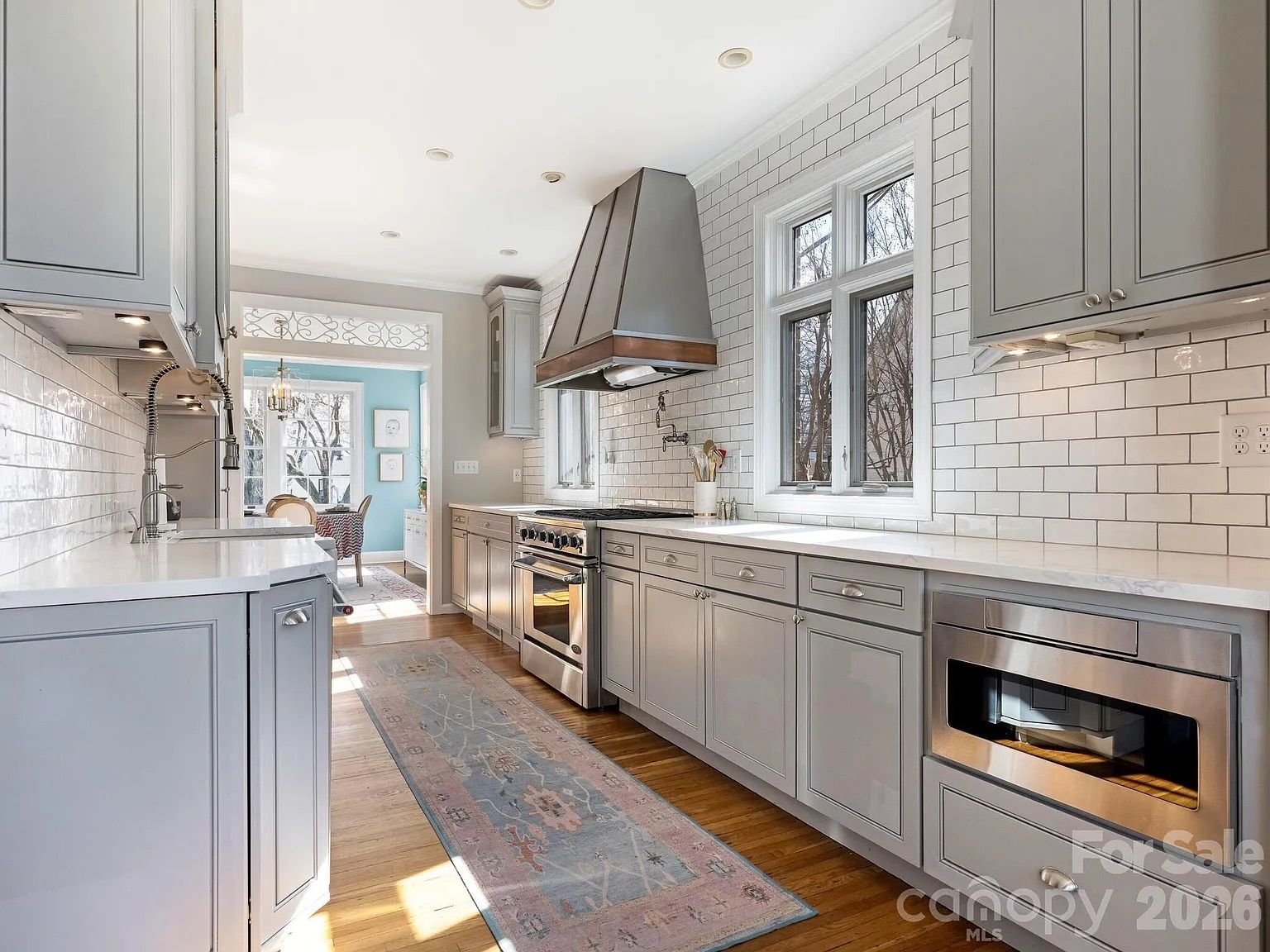 Bright galley kitchen with gray cabinets, white subway tile backsplash, stainless steel stove and microwave, hardwood floors, and a pastel blue and pink runner rug.