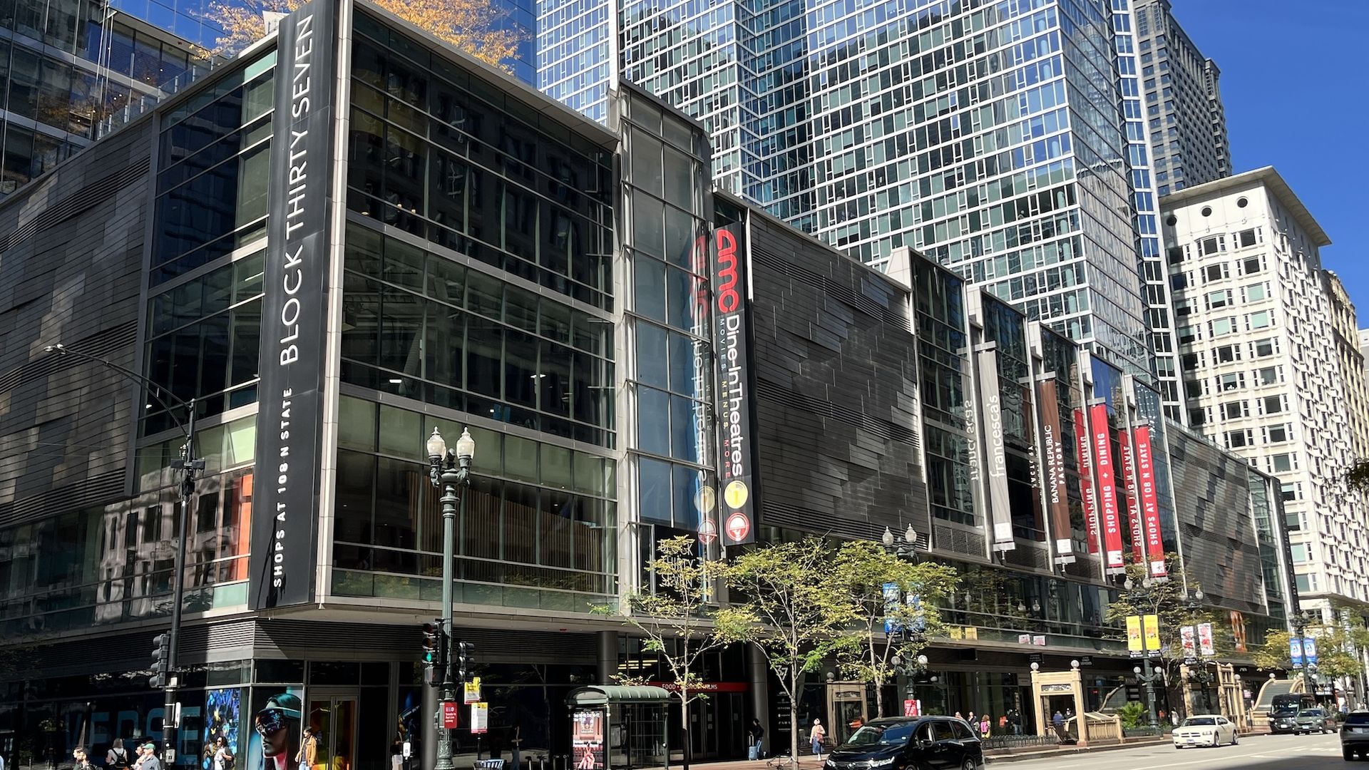 Urban city street with tall glass office towers and blue sky. Colorful banners line the modern building; pedestrians cross the crosswalk as cars drive by and storefronts appear at street level.