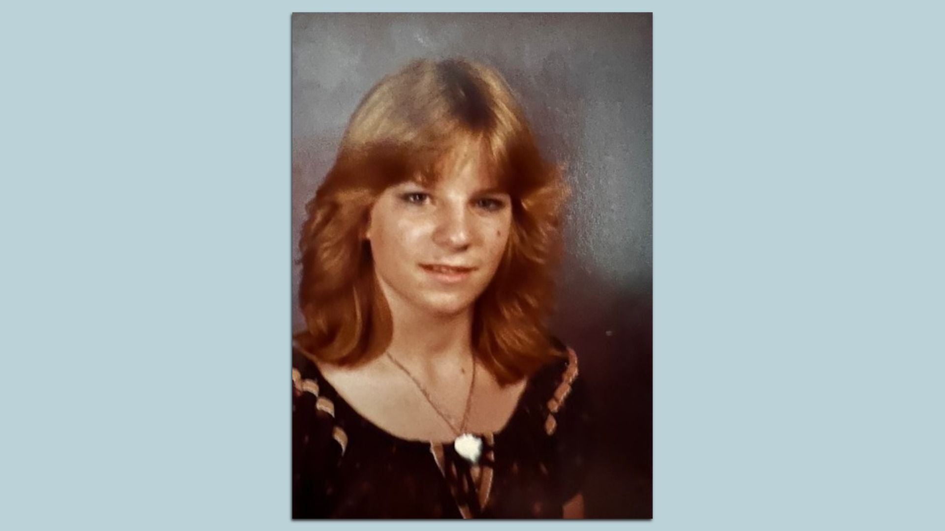 Portrait of a girl with shoulder-length strawberry-blonde hair, wearing a dark floral blouse and a pendant necklace, posed against a textured gray studio backdrop.