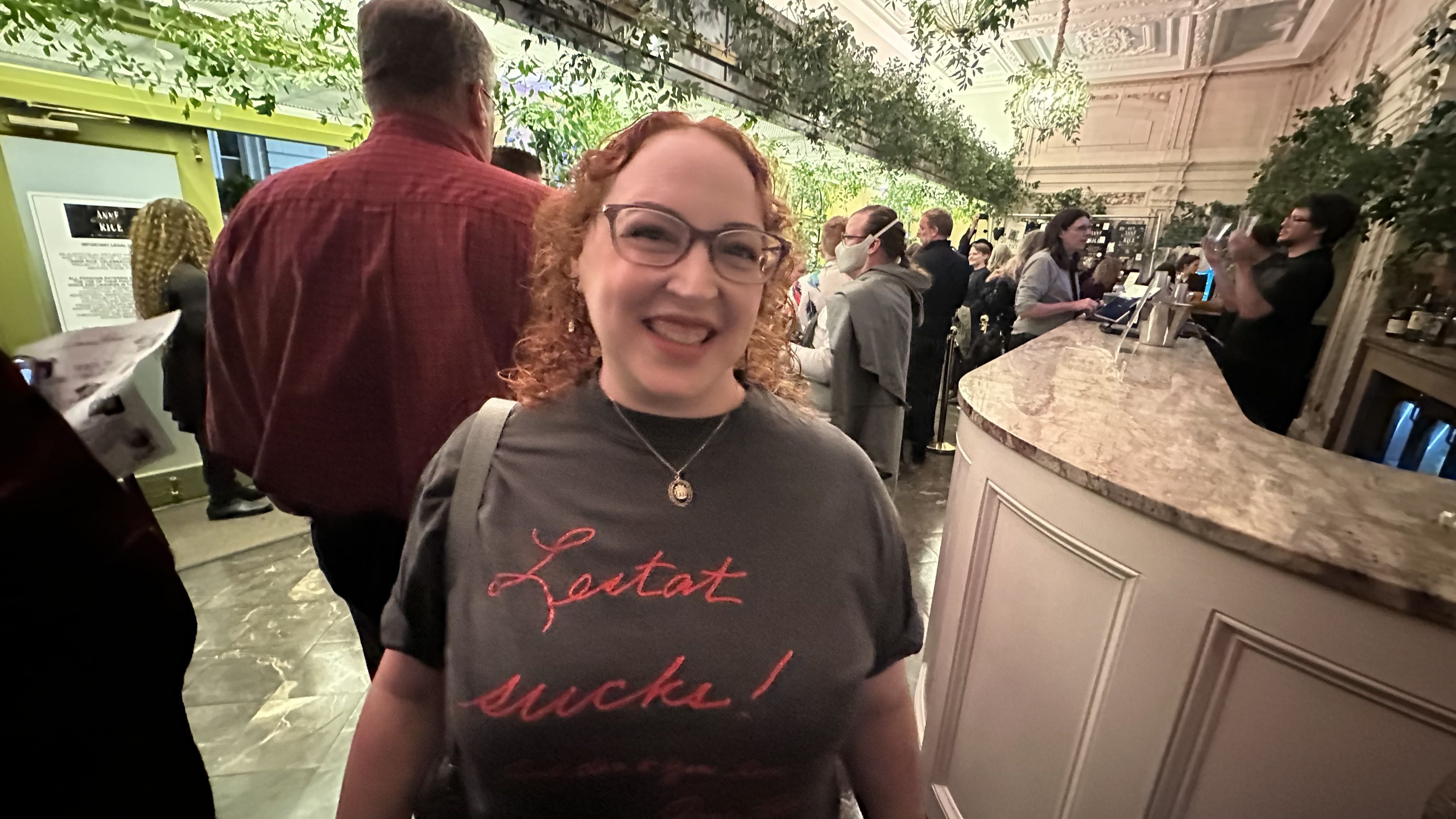Smiling woman with curly red hair and glasses in a crowded, warmly lit venue with green foliage and a marble bar, wearing a grey shirt that says "Lestat sucks!" in red cursive.