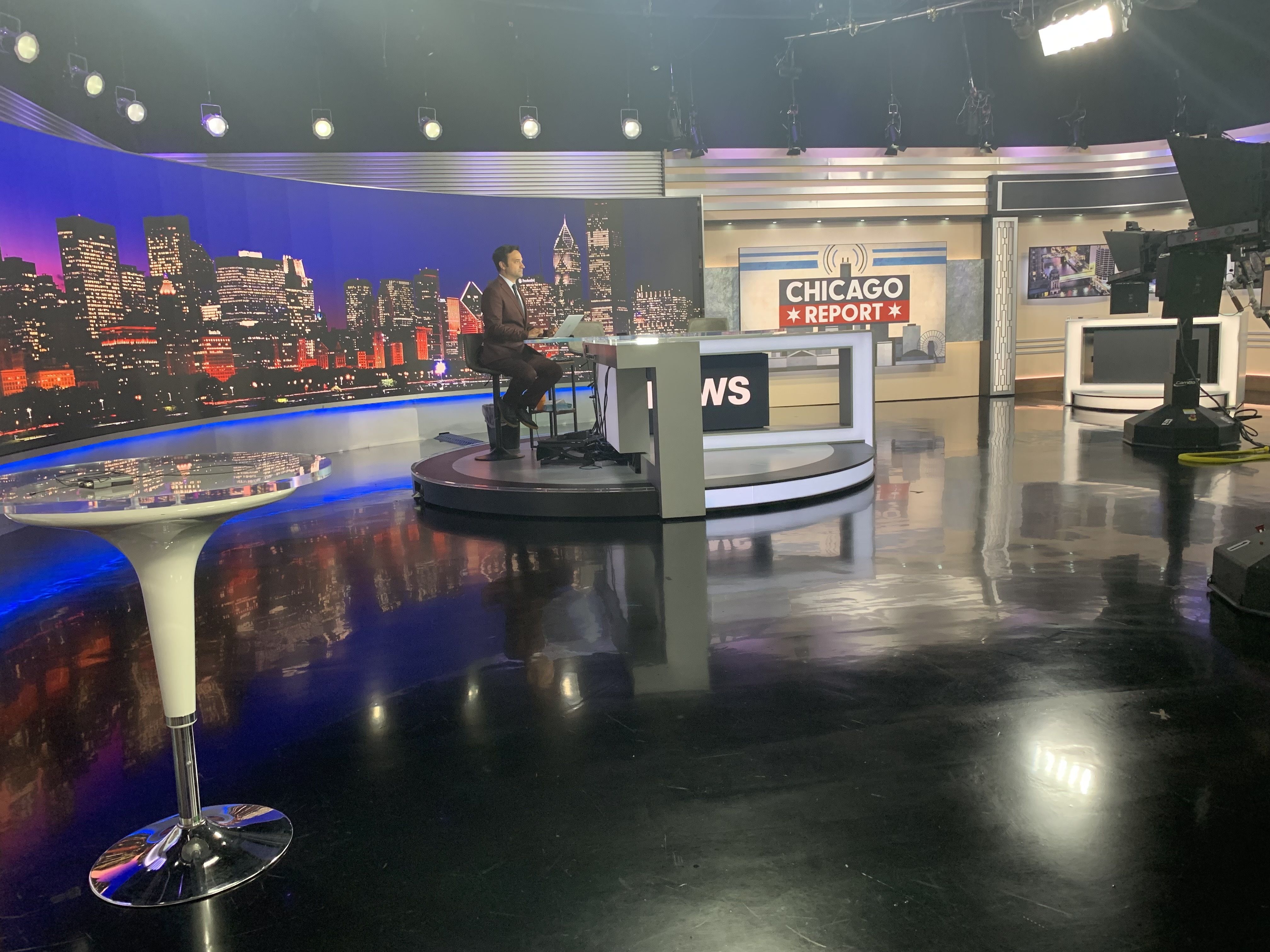 News studio set with a man in a suit at a desk with a laptop, a Chicago skyline backdrop, and a bold "CHICAGO REPORT" sign. Studio lights and camera rigs visible.