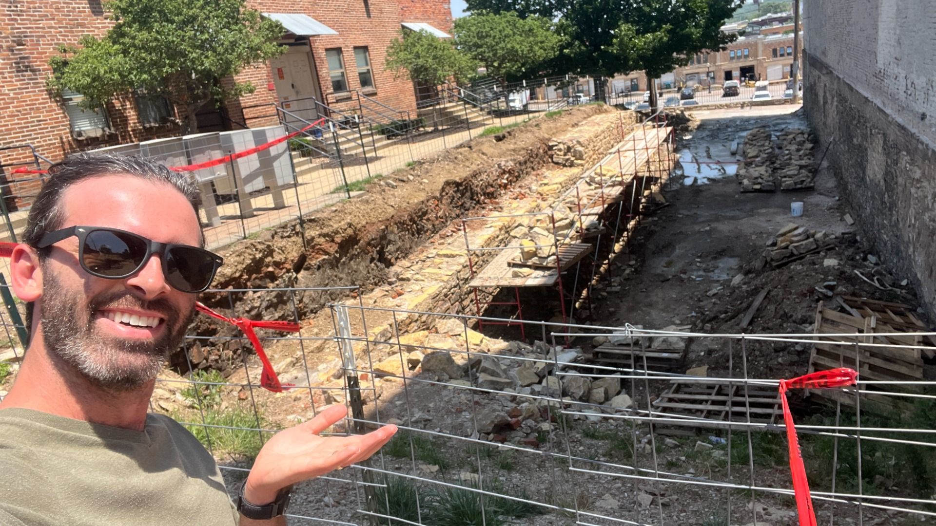 Axios reporter Travis stands in the foreground motioning to a ditch gouged in the earth in between two buildings. Fencing around the pit has red tape tied to it.