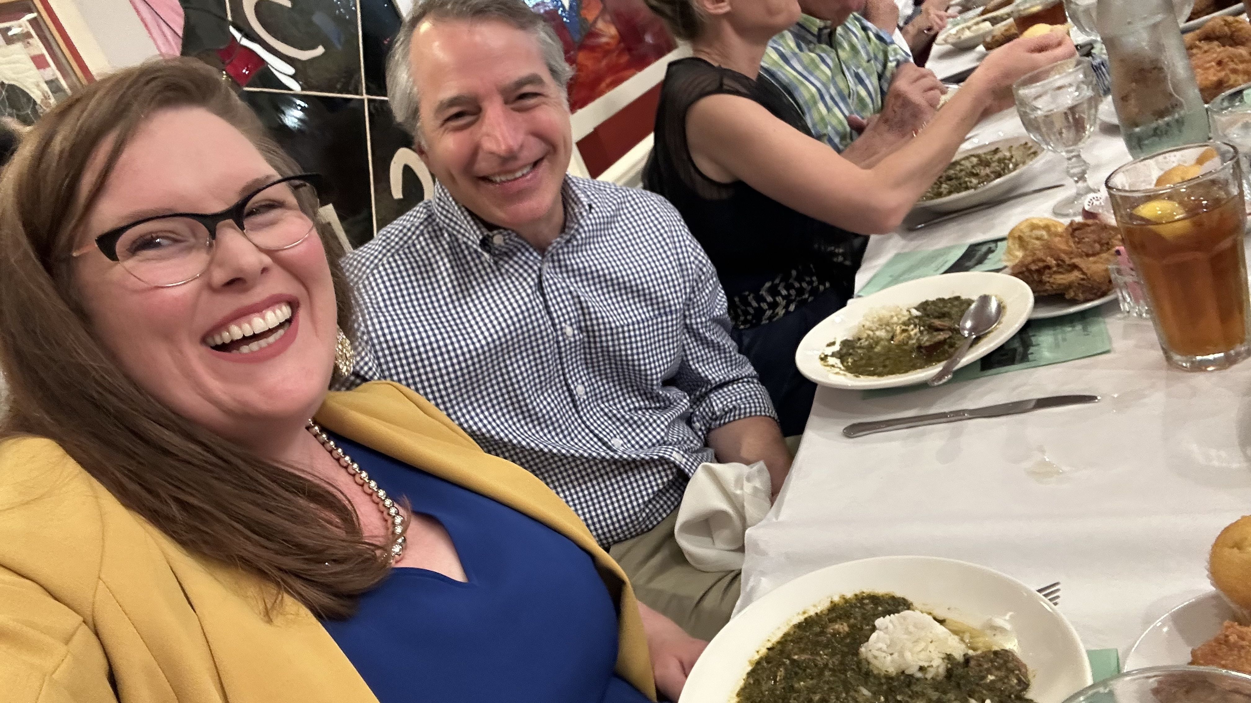 A woman with glasses and a yellow blazer laughs at a long banquet table, seated next to a smiling man in a blue check shirt; diners eat plates of greens, bread, and iced tea.