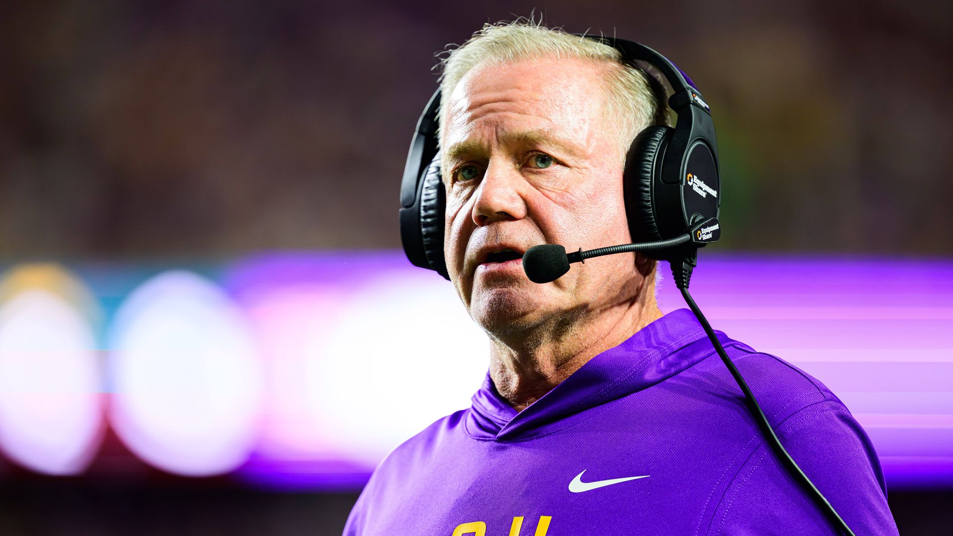 Image show Brian Kelly in an LSU hoodie with a headset on.