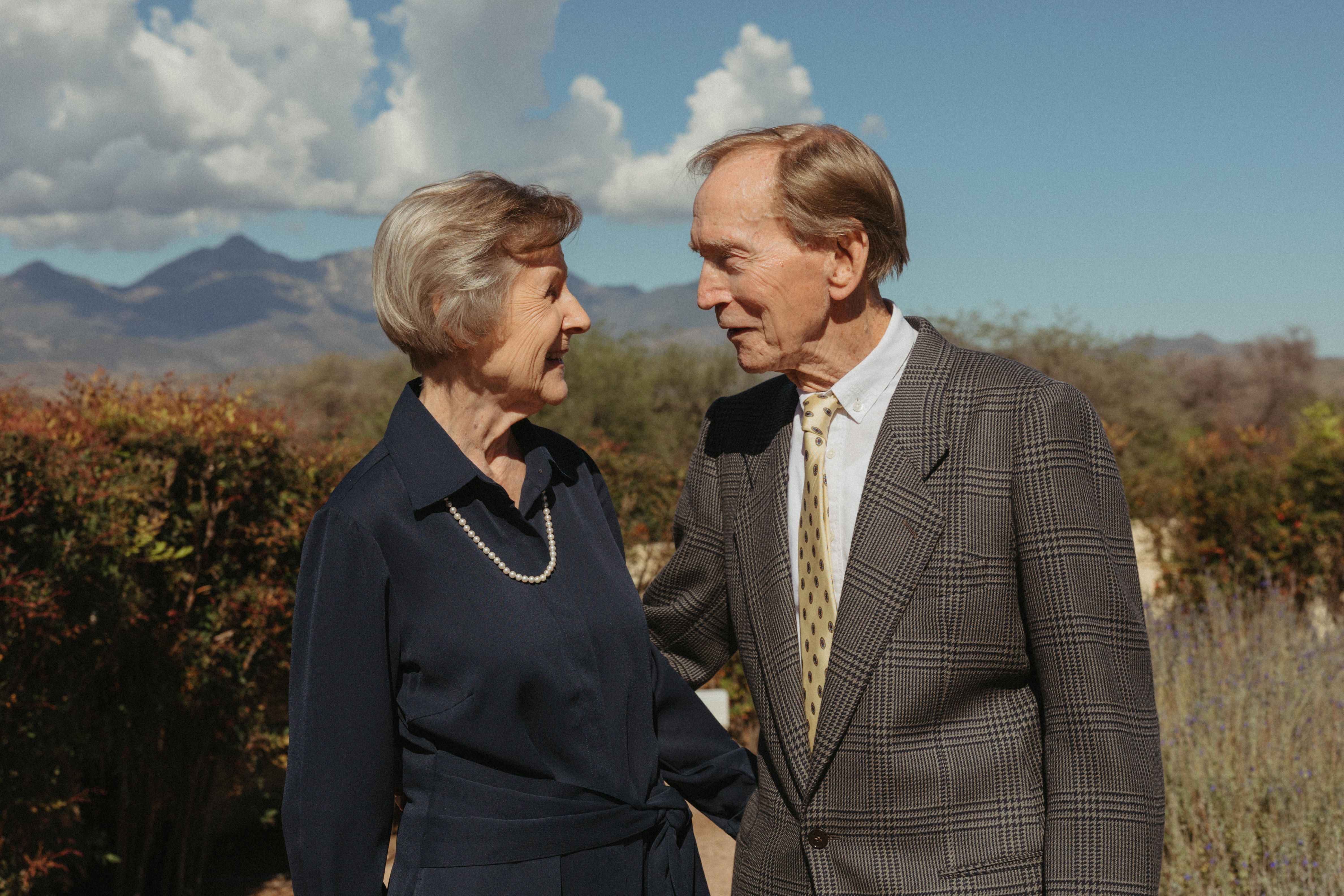 An elderly couple faces each other and smiles outdoors. The woman wears a navy dress with a pearl necklace; the man wears a plaid suit and tie, with mountains and a blue sky behind.