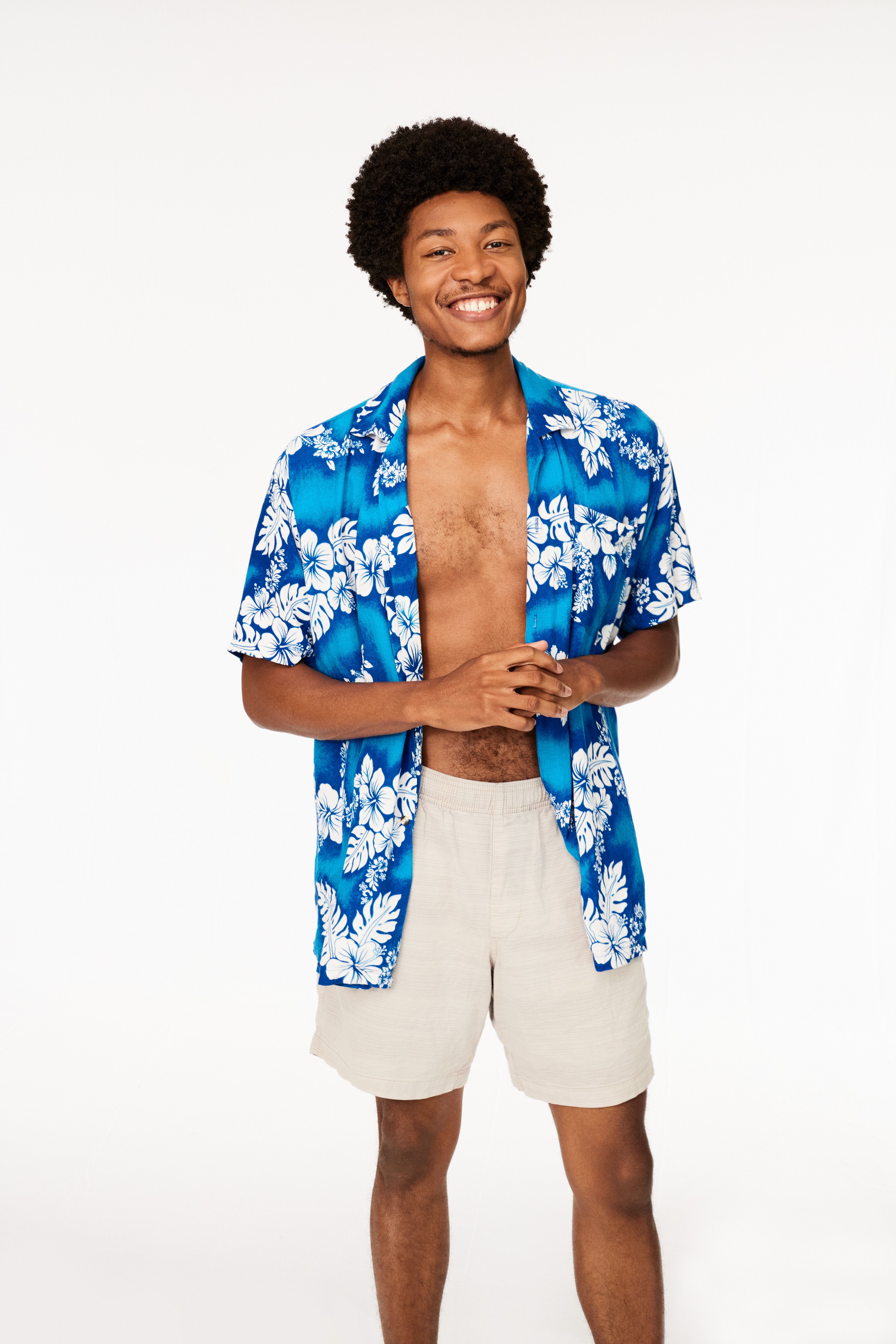 Smiling young man with curly hair wearing an open blue Hawaiian shirt with white floral patterns and beige shorts, standing against a white background.