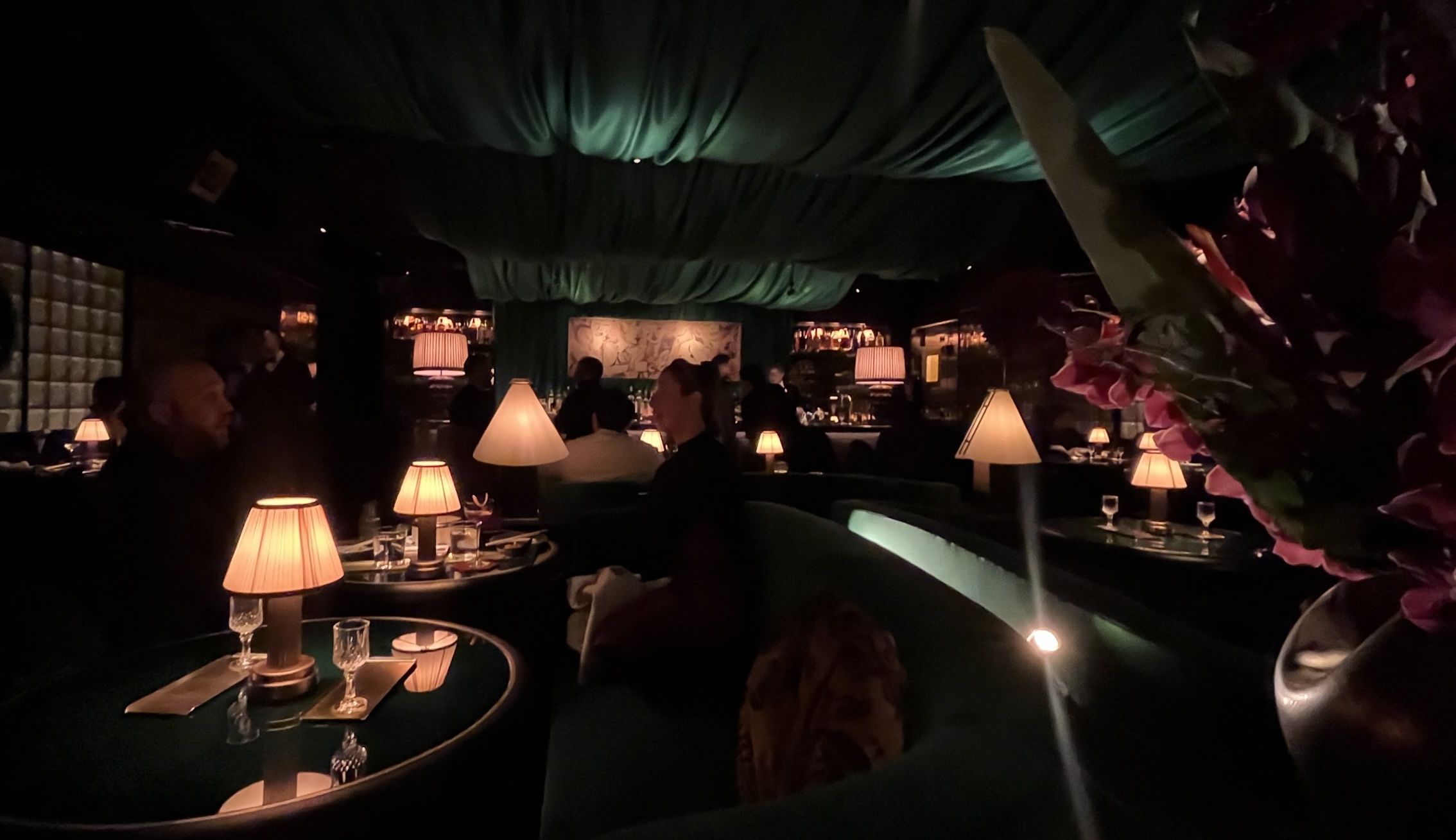 Dim upscale restaurant with a green draped ceiling, warm table lamps, round glass tables, and diners. A bar in the back, plush seating, and a large pink flower arrangement on the right.