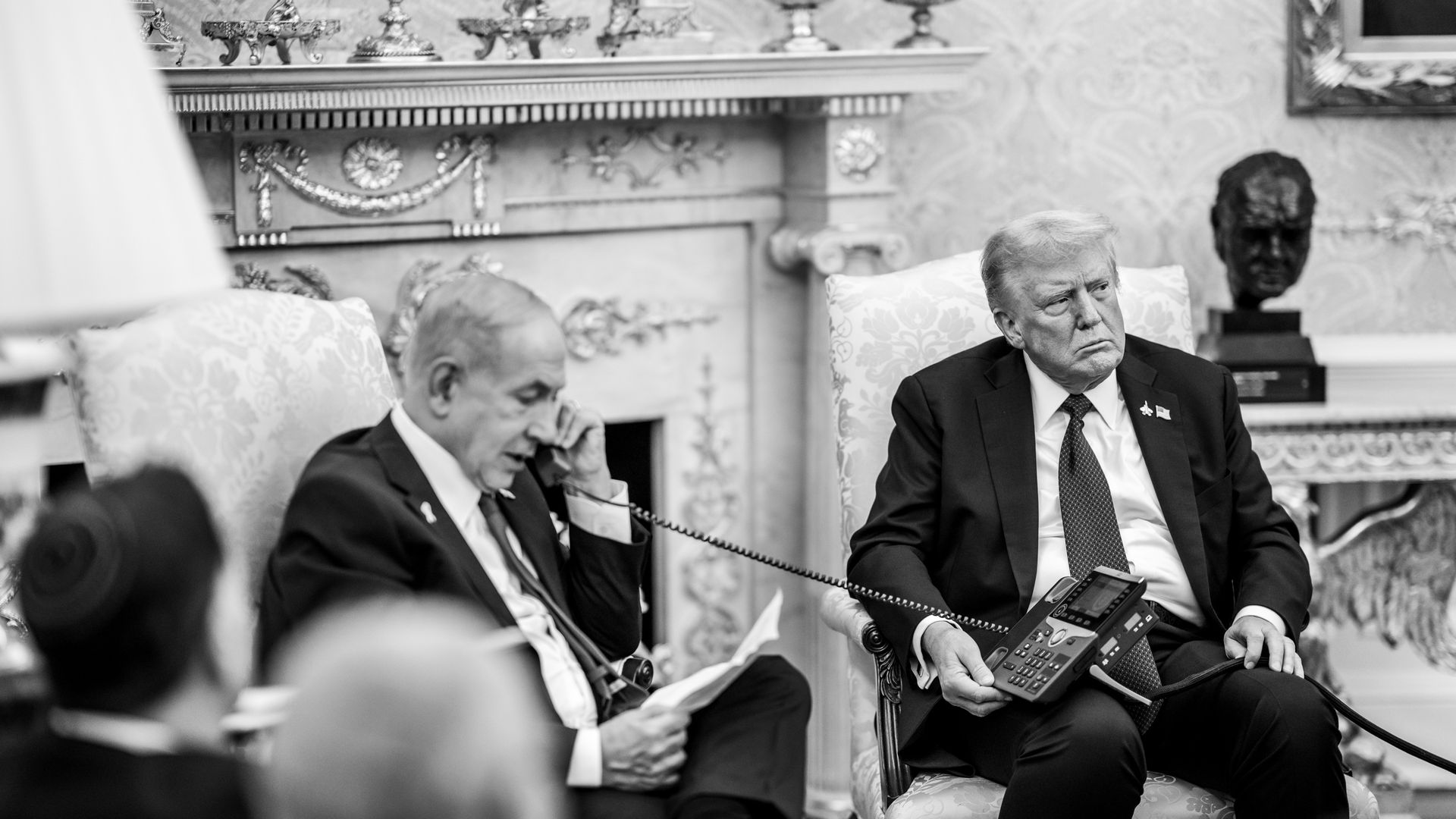 Black and white photo of two men in suits seated in a formal room. One holds a phone receiver and paper; the other holds the phone base, looking serious. Ornate fireplace and bust in background.