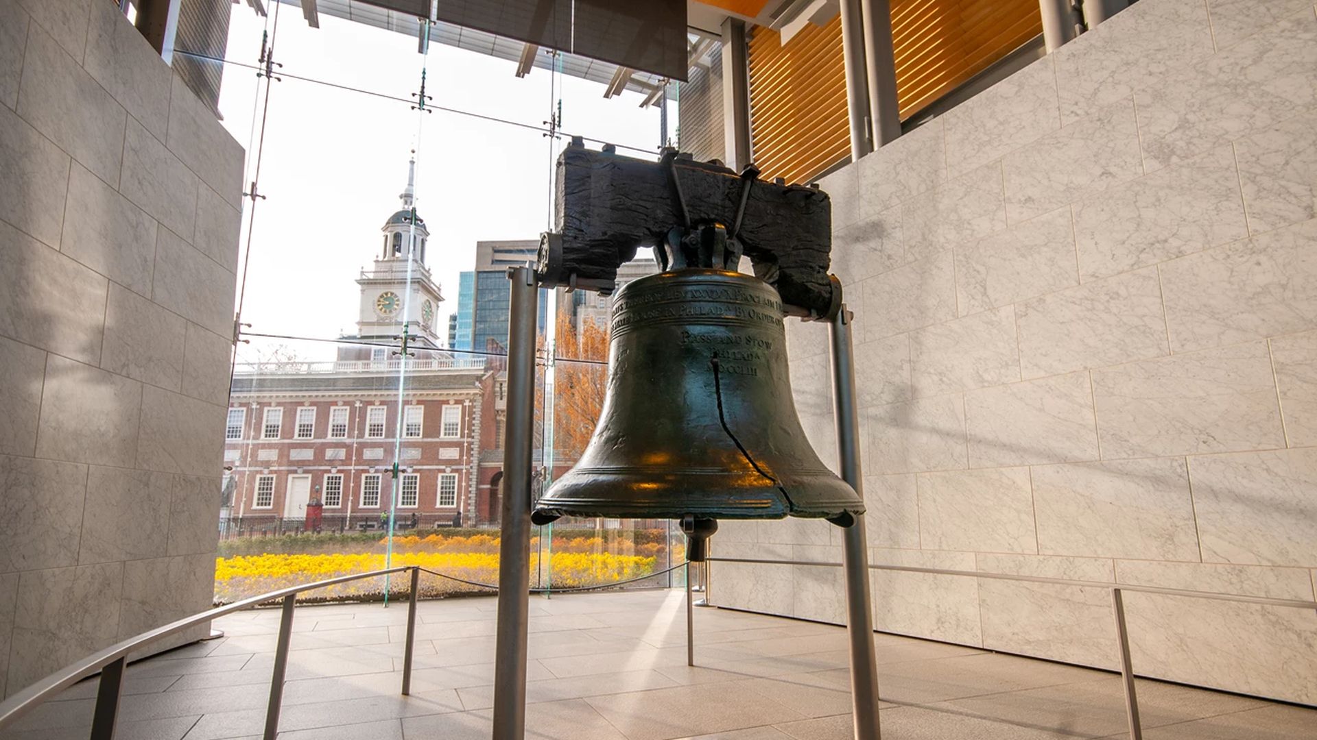 The Liberty Bell with its iconic crack displayed inside a modern building with glass walls and marble panels, with Independence Hall visible outside in the background.
