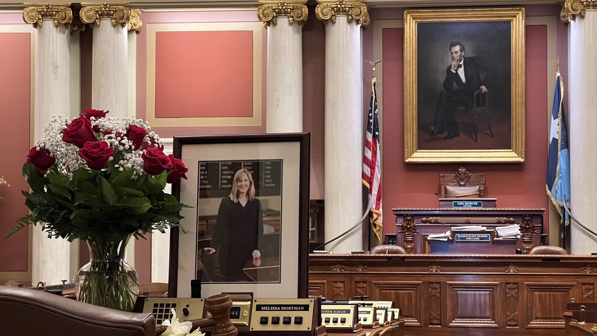 The Minnesota House of Representatives chamber with a large bouquet of red roses, a framed photo of a woman in a black suit, wooden desks with nameplates, marble pillars, and a portrait of Abraham Lincoln.