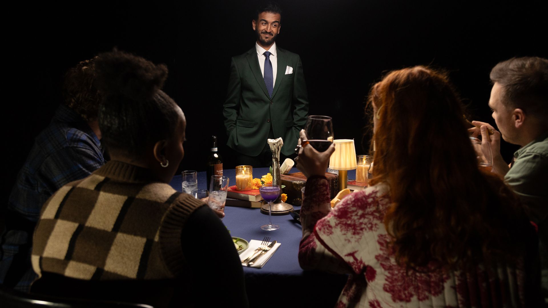 A man in a dark green suit stands at a dining table, smiling, as guests sit around. The table has candles, wine glasses, a beer bottle, and a blue tablecloth.