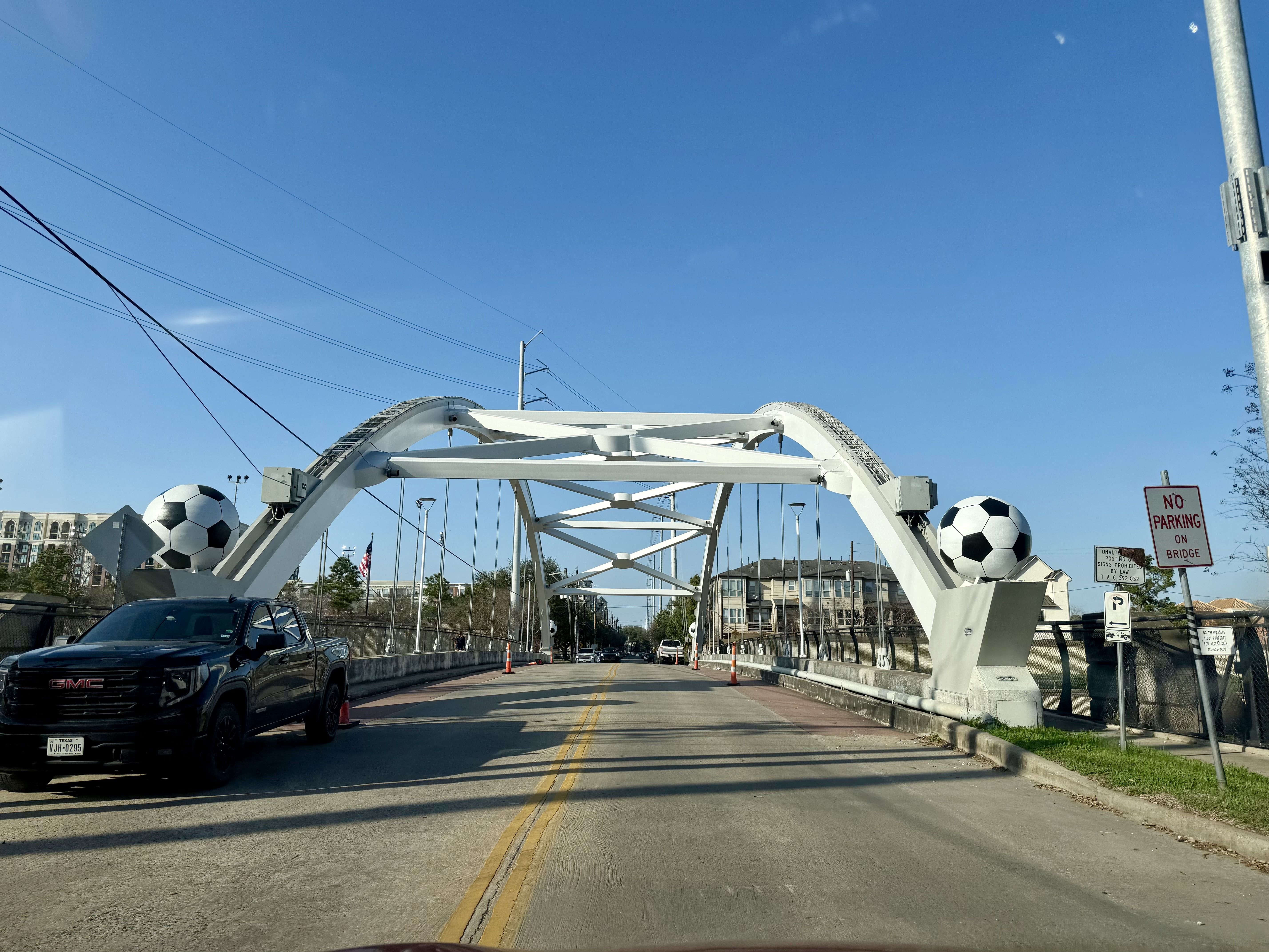 White arch bridge with large soccer-ball sculptures on both sides, cars on the road, overhead power lines, bright blue sky, and a 'No Parking on Bridge' sign on the right.
