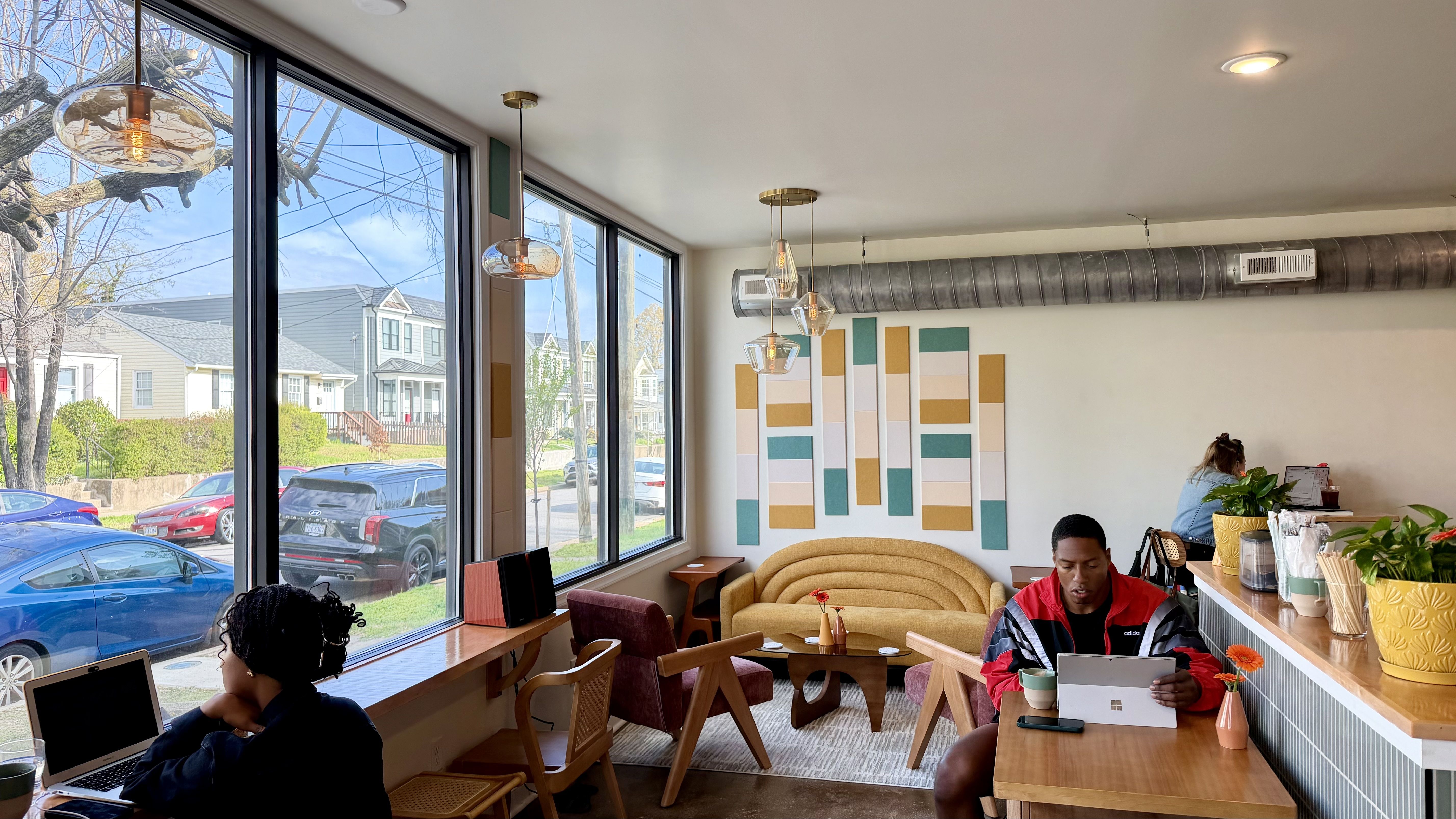 Bright cafe with large windows overlooking a street of houses. Beige curved sofa, wooden chairs, and a coffee table. A man in a red jacket uses a tablet; a woman works on a laptop. Potted plants and colorful wall art.