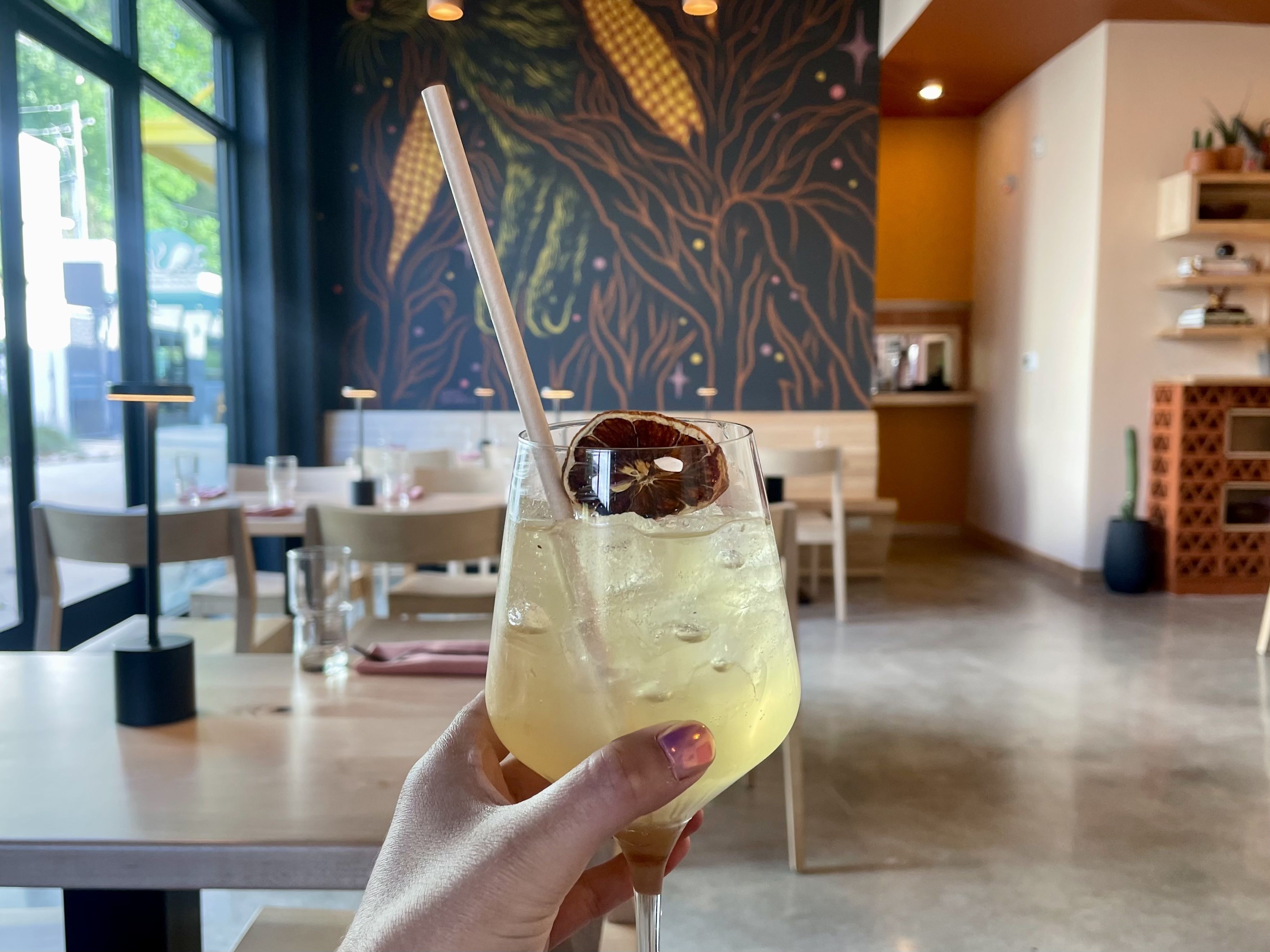 Hand holding a wine glass with a light yellow iced drink garnished with a dried citrus slice and a paper straw, inside a bright, modern cafe with colorful wall art and wooden furniture.