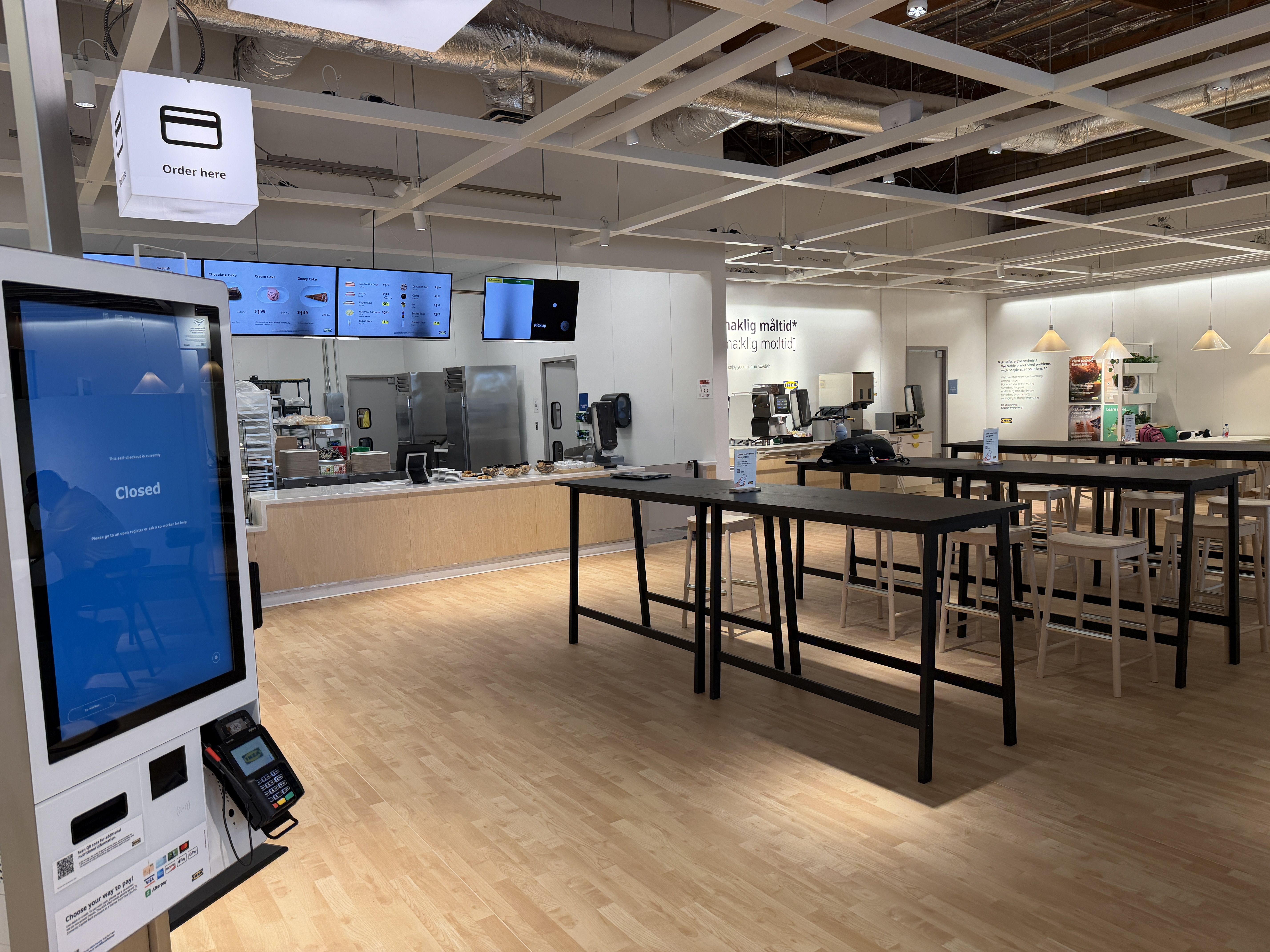 Bright cafeteria with a service counter and menu boards. A blue 'Closed' kiosk on the left, white high tables with stools, and an 'Order here' sign overhead; exposed ceiling pipes.