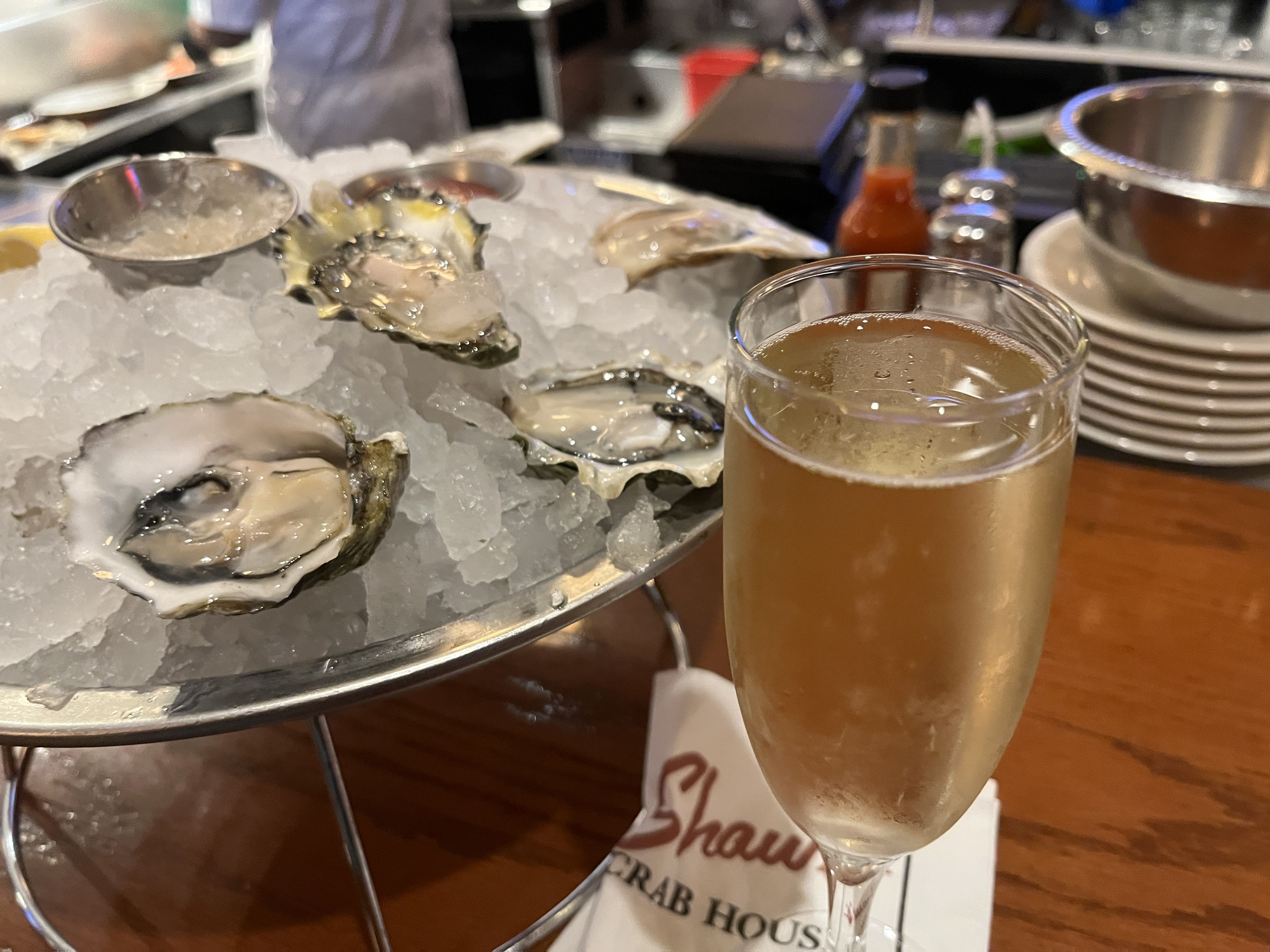 Plate of oysters on crushed ice, served with sauces in metal cups, next to a glass of sparkling white wine on a wooden table at a seafood restaurant.
