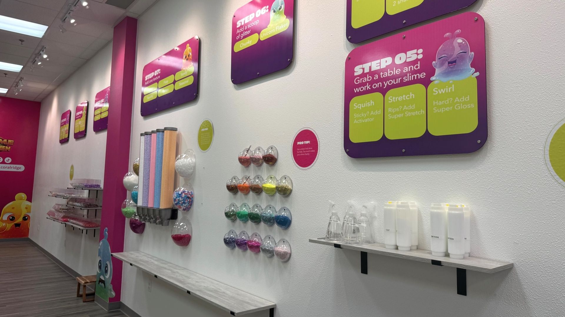 Interior of a brightly lit slime workshop with colorful signs on white and pink walls detailing steps for making slime and shelves holding various slime ingredients and tubes.