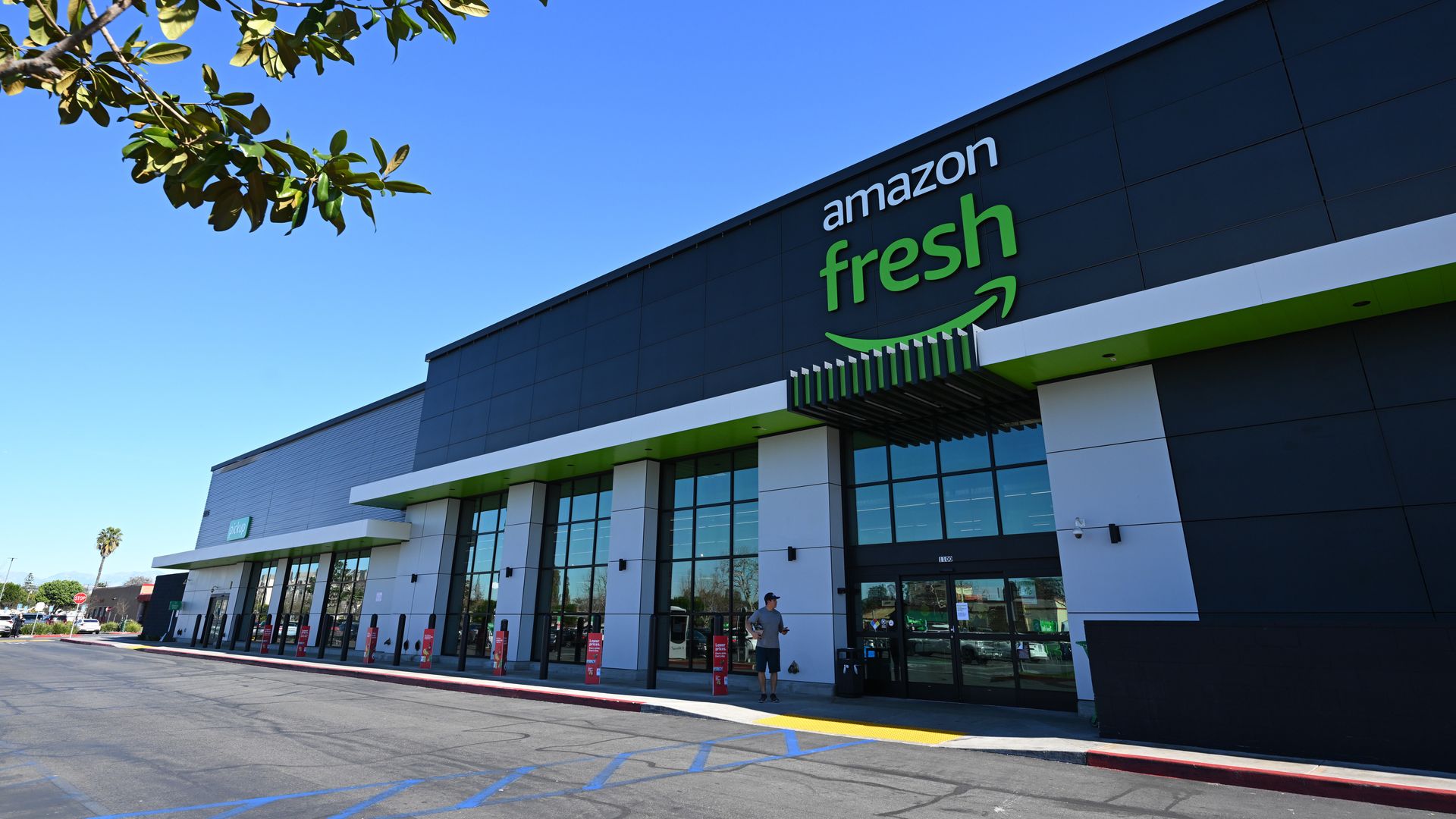 Exterior view of an Amazon Fresh grocery store with black and white facade, green accents, and clear skies with tree branches in the foreground.