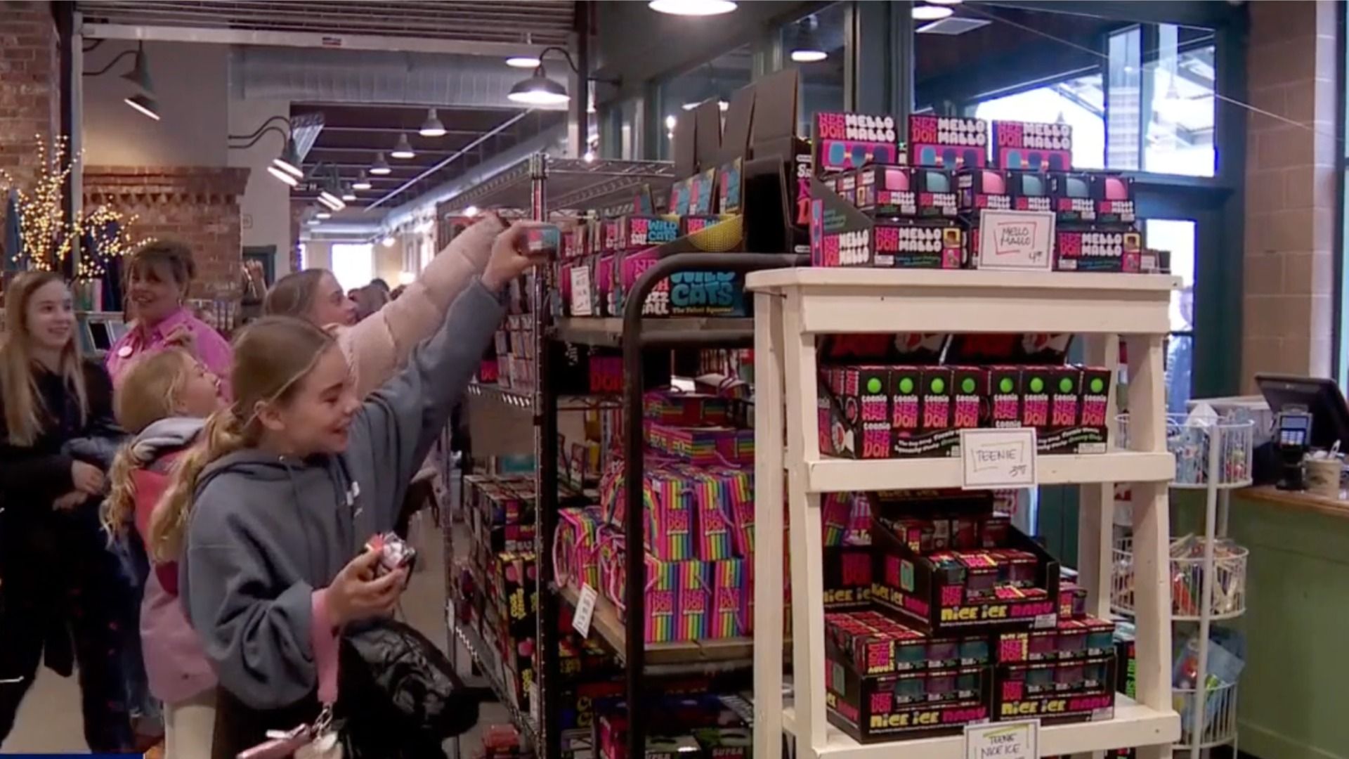 Inside a colorful toy shop, a girl in a gray hoodie reaches up to grab a boxed item from a shelf full of bright, neon packaging while several other kids stand nearby and watch.