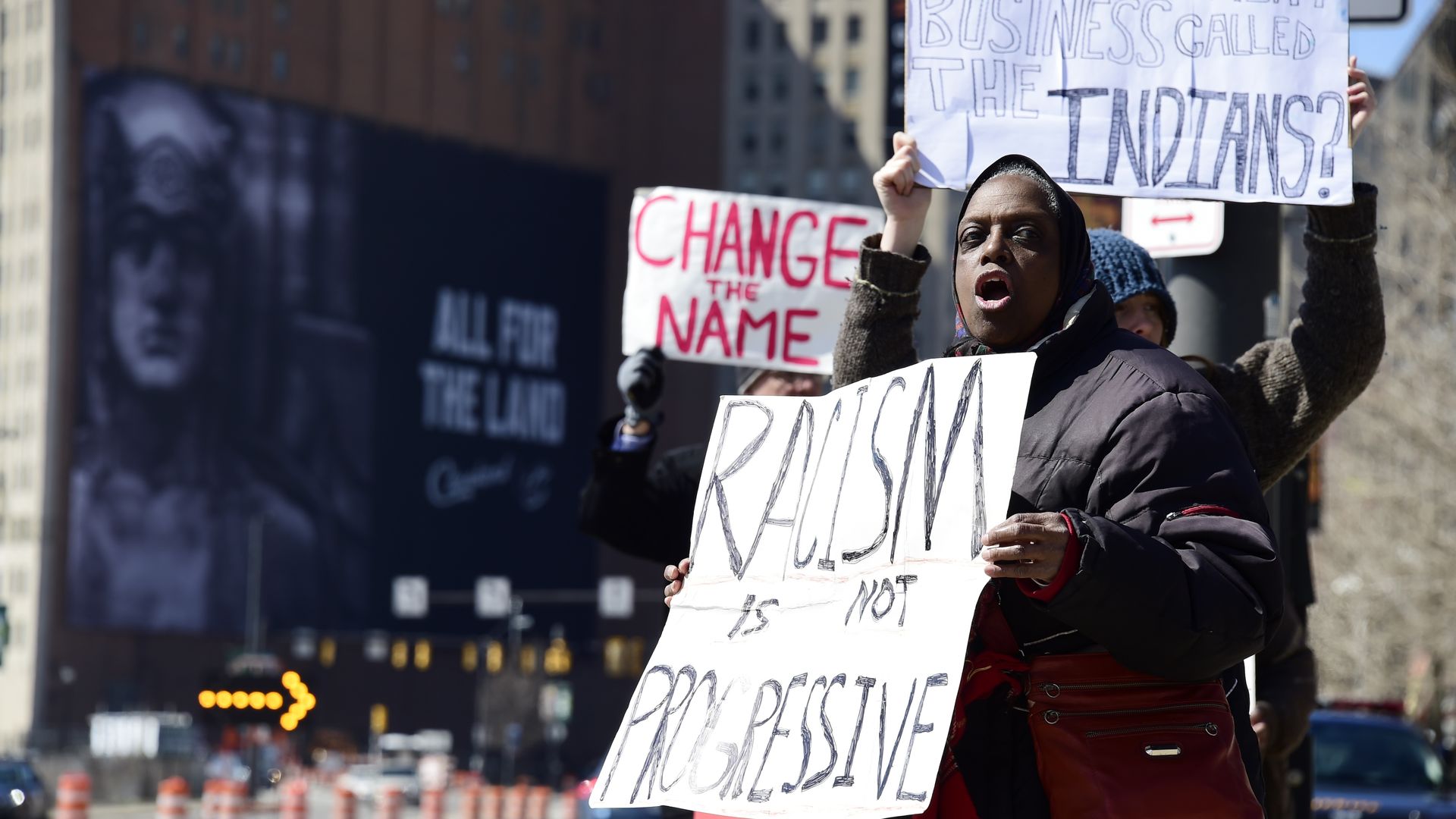 A group of protesters on a city street holding signs, including one that says "RACISM is NOT PROGRESSIVE" and another partially visible saying "CHANGE THE NAME".