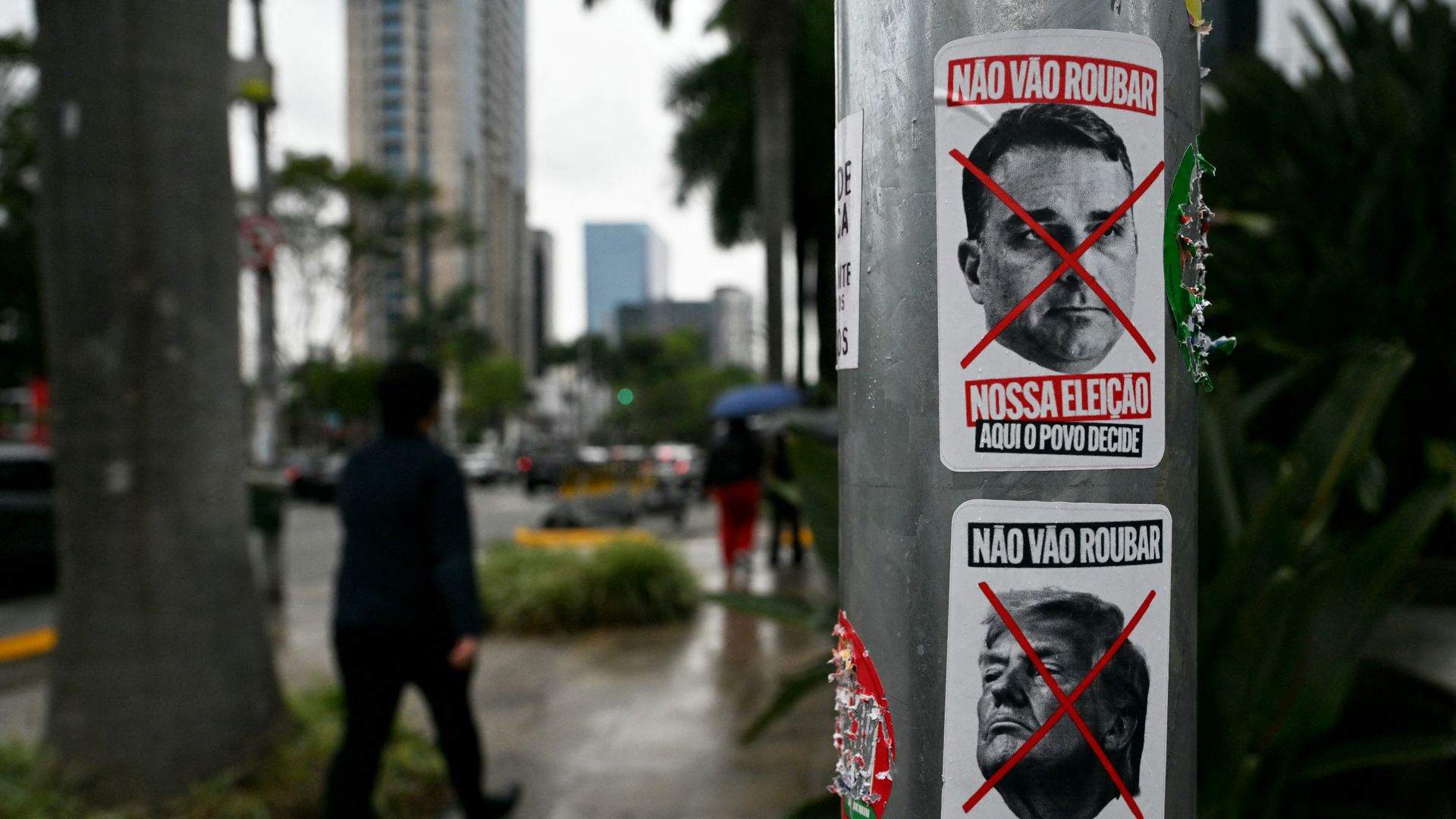Two white posters with men's faces (on top, Flavio Bolsonaro, and beneath, President Trump) crossed out with red Xs appear on a silver utility poll as people walk past on a sidewalk.