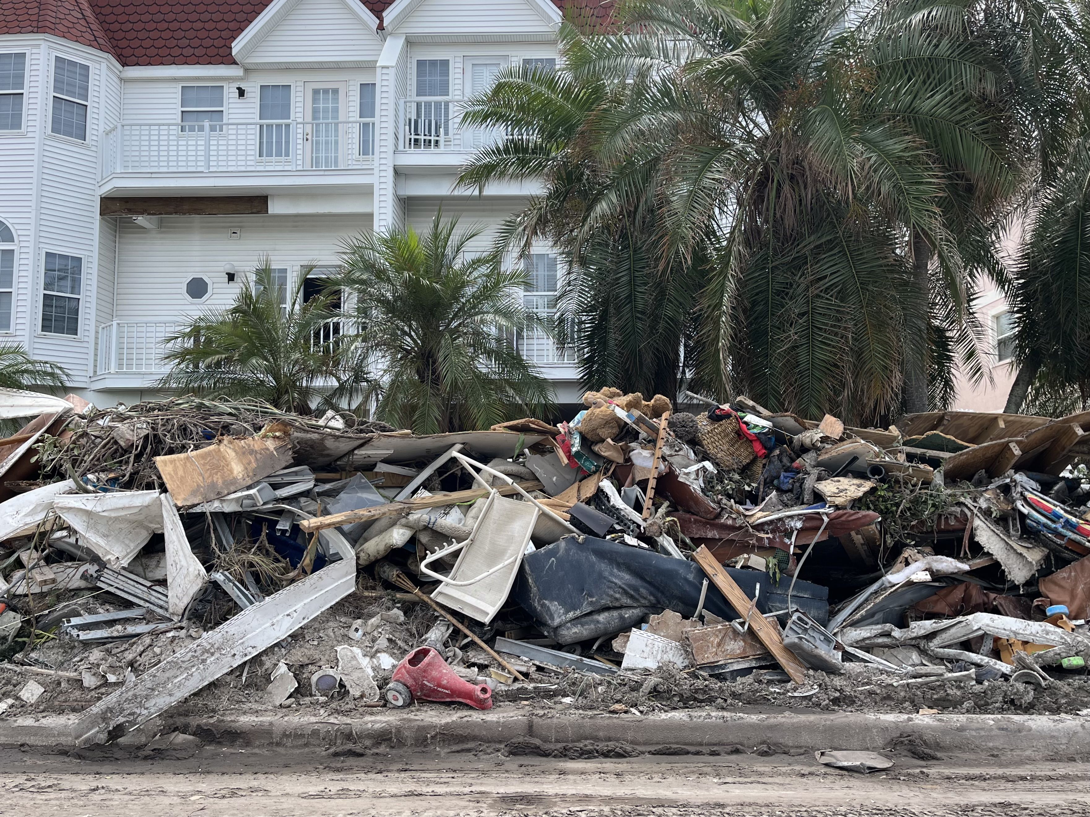 A pile of debris, window frames, furniture and more piled in front of a white apartment building with palm trees in front of it.