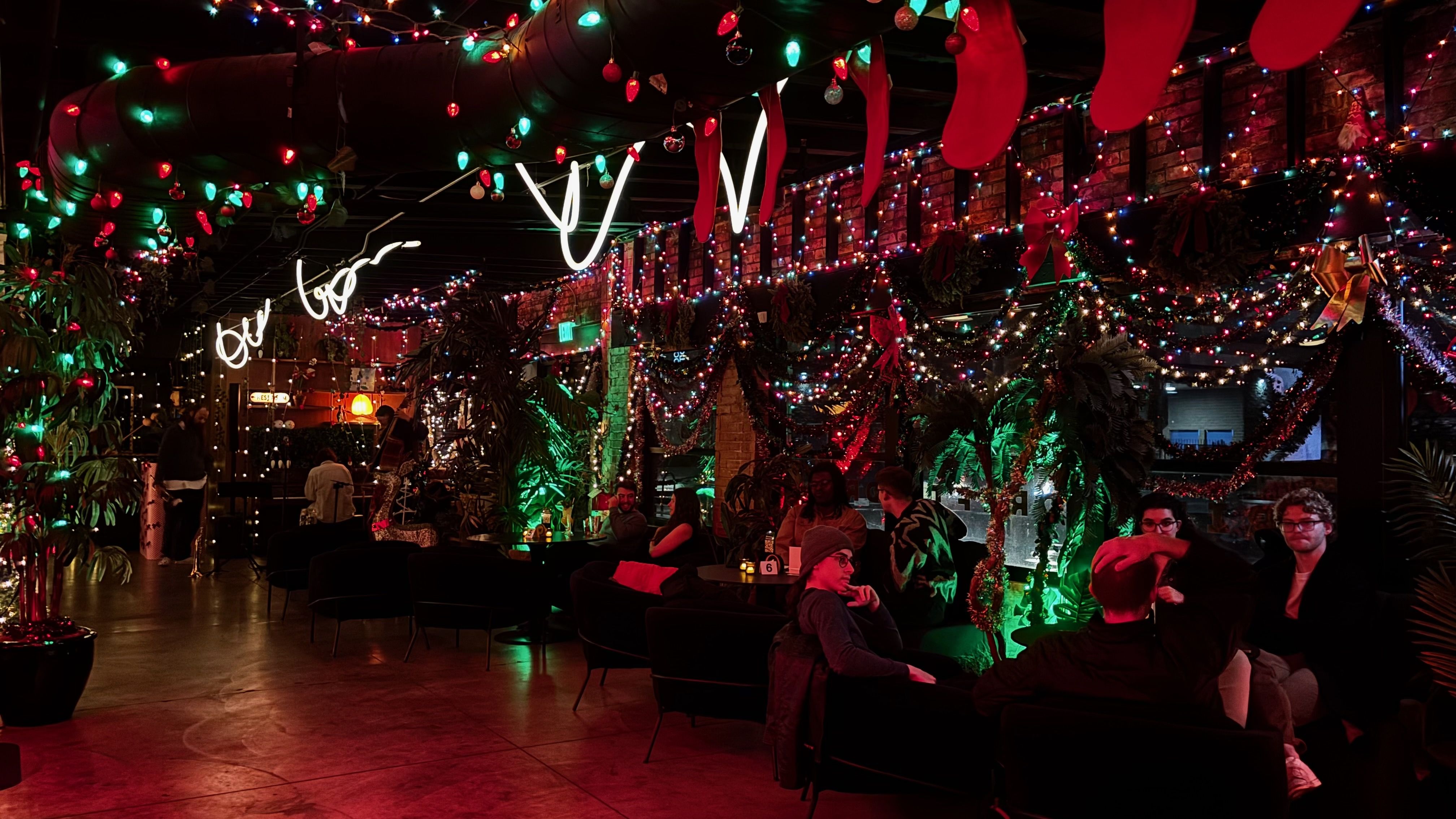 Indoor space decorated with colorful Christmas lights, red stockings, wreaths, and garlands; people seated on black chairs and sofas, chatting in a festive, dimly lit atmosphere.