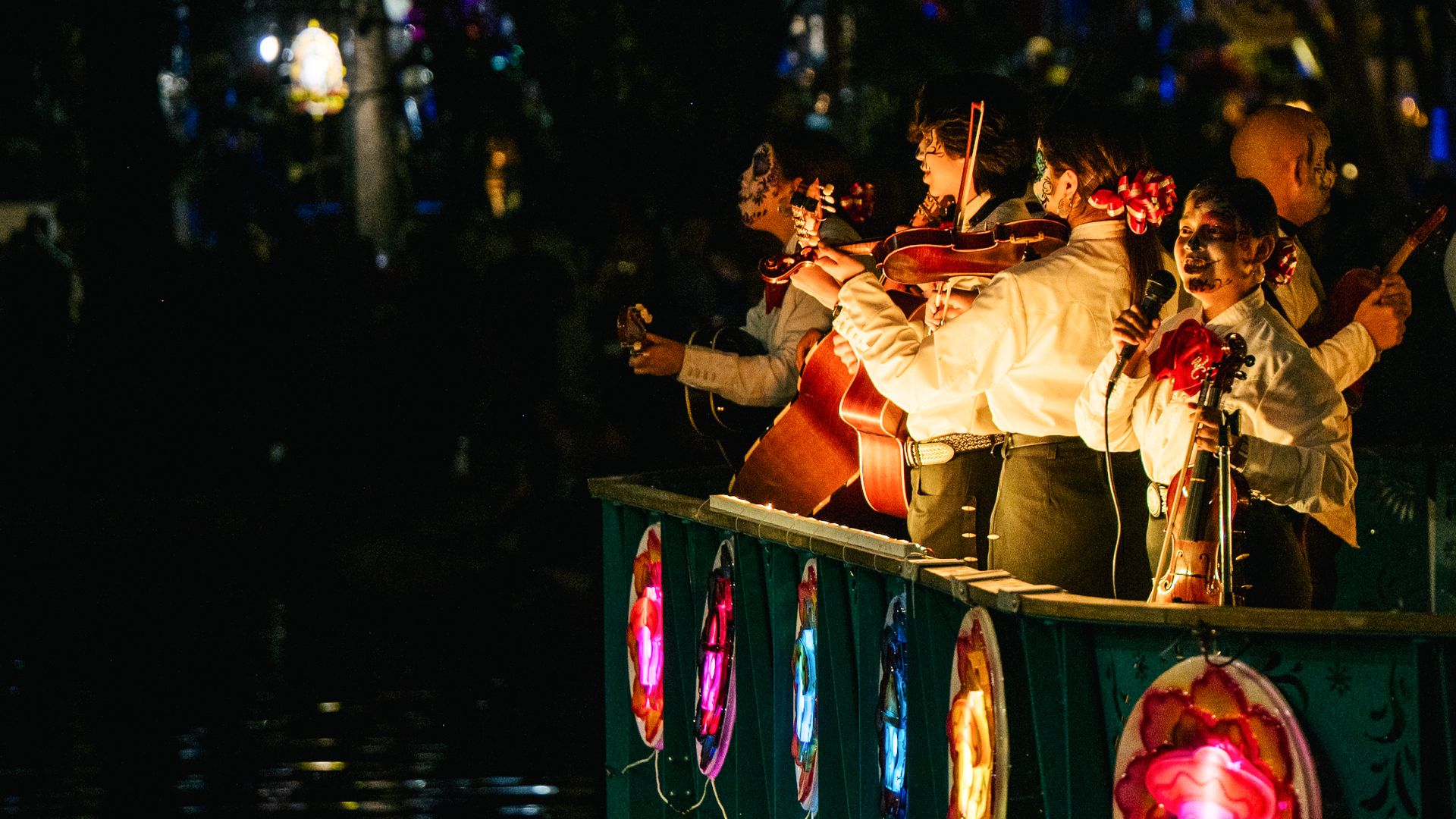 A Mariachi band plays aboard a cruise ship float during the Day of the Dead River Parade on the San Antonio River in 2021. The musicians are wearing skeleton face paint and flowers on their shirts and in their hair, and are lit up by lights against a dark background.