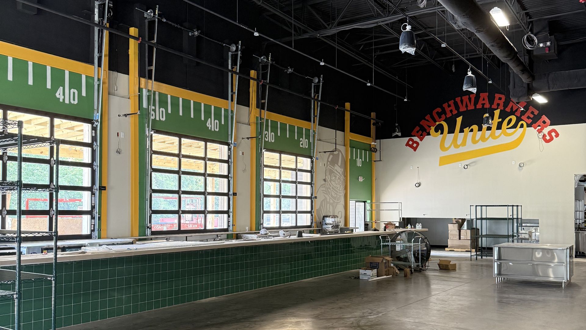 Interior of a sports-themed restaurant or bar under construction with green tile counter, football field markings on walls, large windows, and "Benchwarmers Unite" mural in red and yellow.