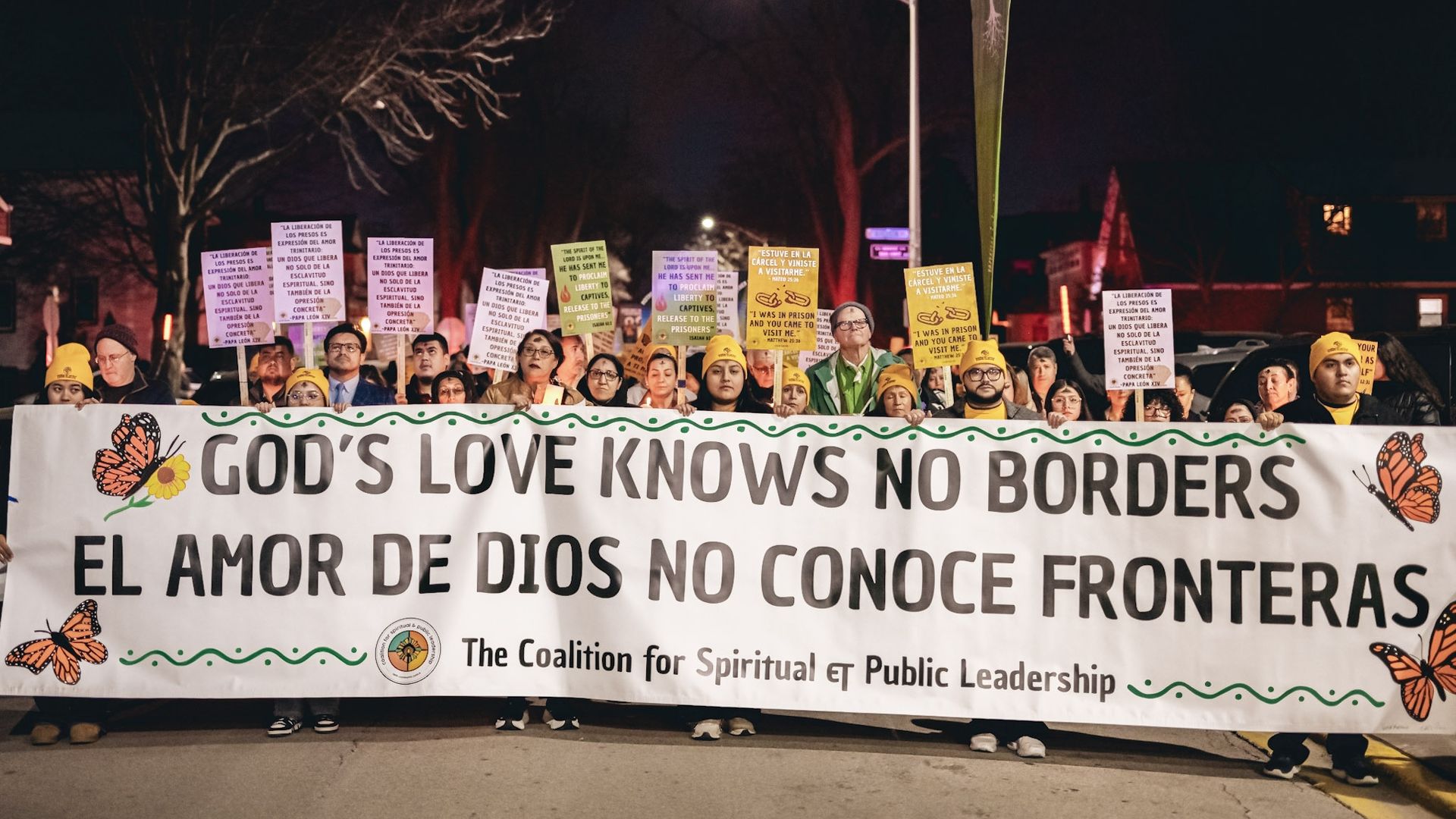 Nighttime protest with a long banner reading GOD'S LOVE KNOWS NO BORDERS. Diverse crowd in yellow hats holds placards; butterflies decorate the banner, and a streetlamp glows in the background.