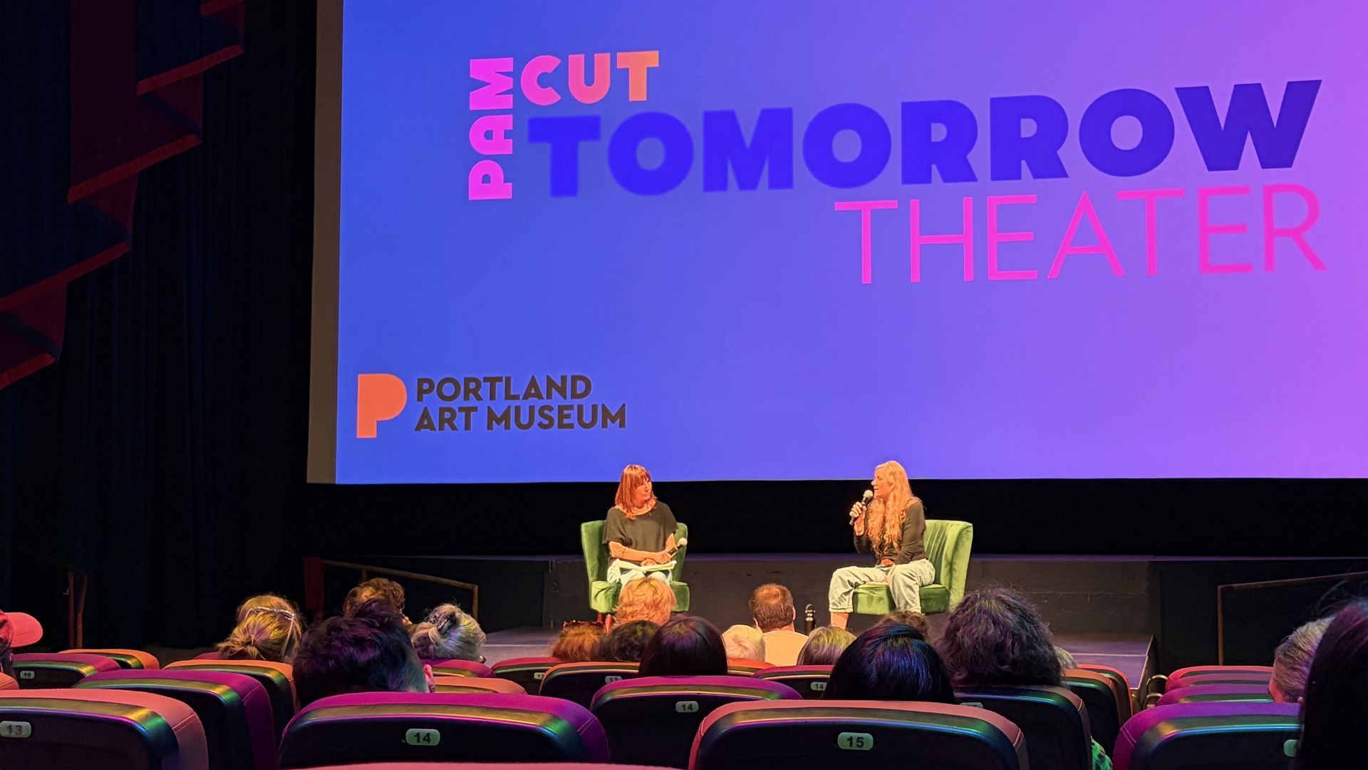 Stage talk at the Portland Art Museum: two women in green chairs on a dark stage, a purple-blue gradient screen behind them reading "TOMORROW THEATER"; audience visible in front.