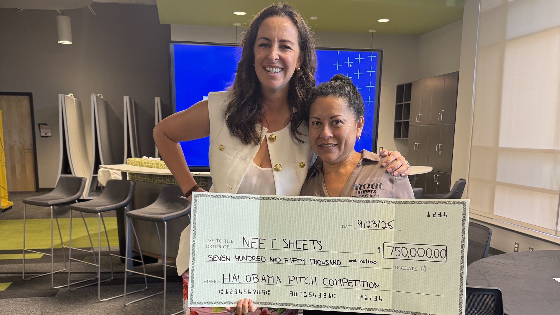 Two smiling women holding a large check for $750,000 made out to NEET Sheets from the Halobama Pitch Competition, standing in a modern office with gray and green decor.