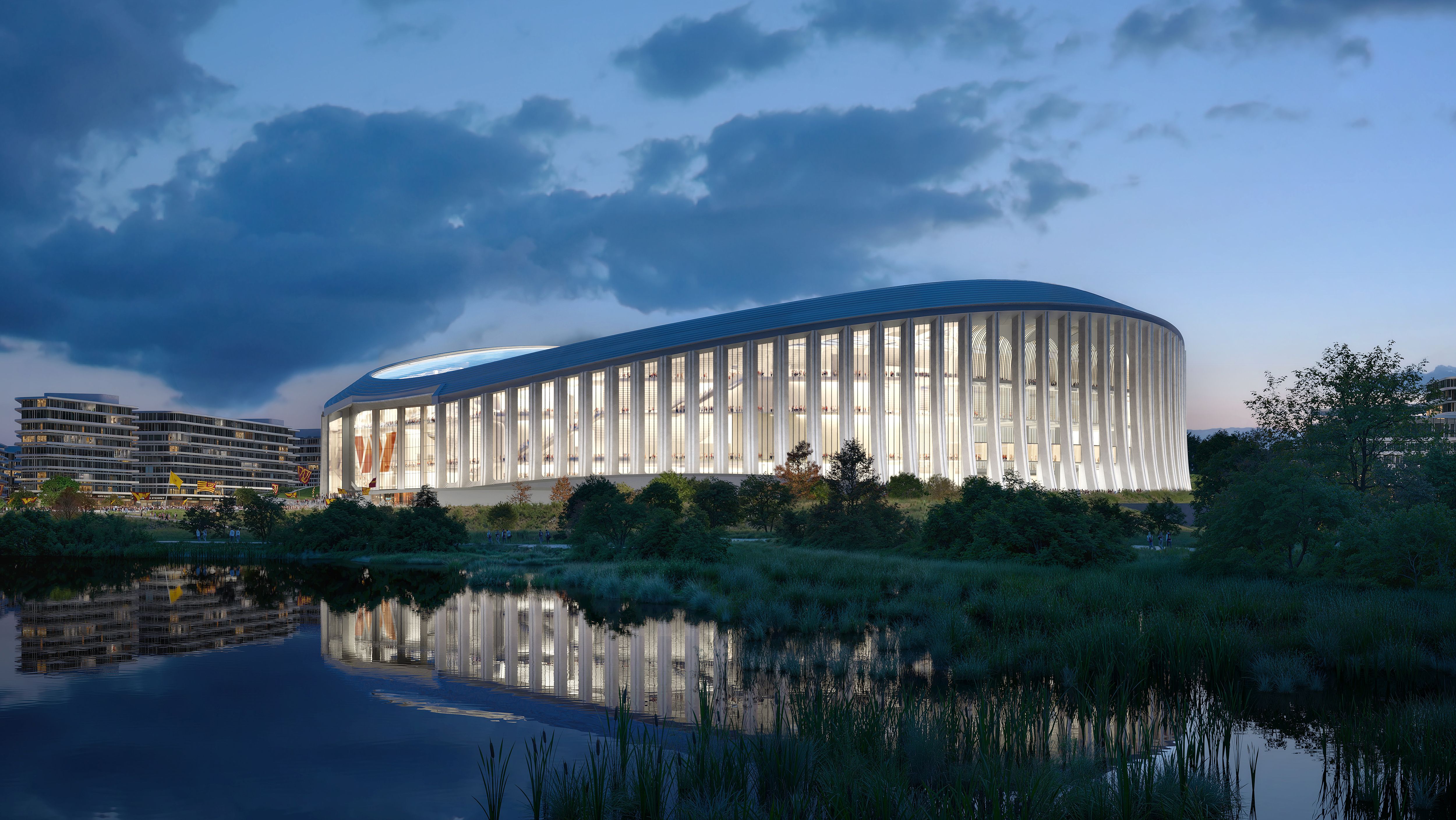 Modern illuminated stadium with vertical white pillars reflecting in a pond, surrounded by greenery and nearby contemporary buildings under a cloudy evening sky.