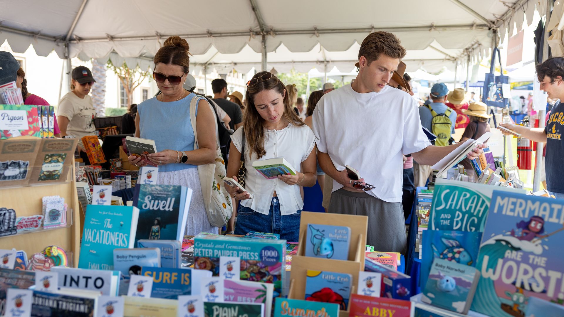 Outdoor book fair under a white tent. Foreground trio browses colorful book tables: a woman in a blue sleeveless dress with sunglasses, a young man in a white shirt, and a girl in white reading.