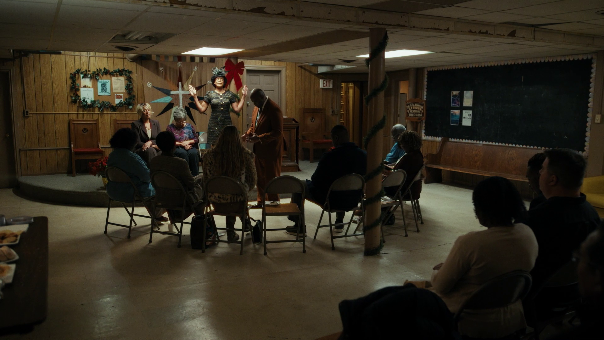 A small, wood-paneled room hosts a community performance. A woman in a glittery dress raises her arms beside a man in a brown suit; folding chairs form a semicircle of spectators, festive decor around.