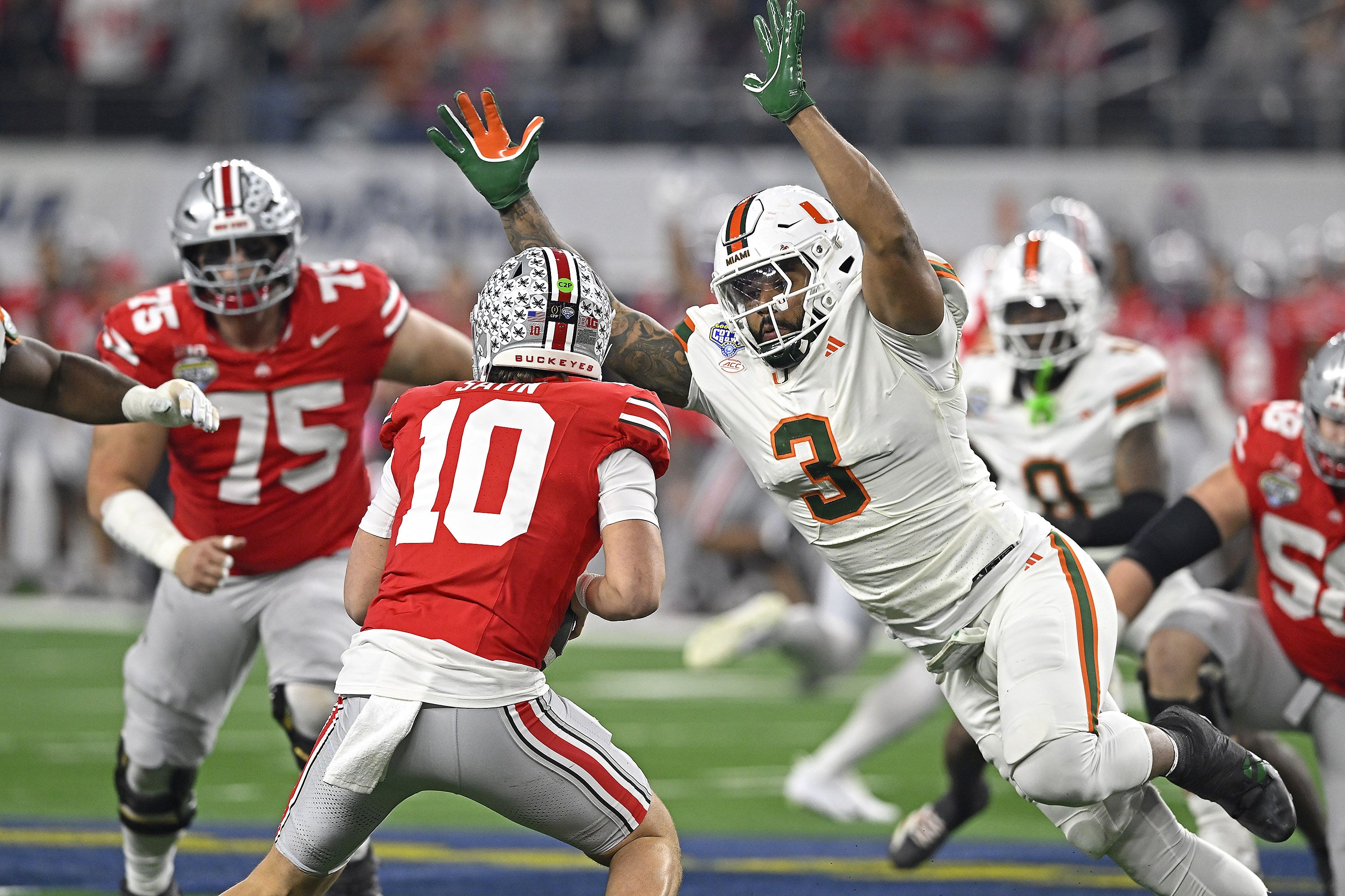 Intense football: Miami Hurricanes player in white jersey leaps to block a Buckeyes pass, facing a red Ohio State player wearing number 10 on a green field.