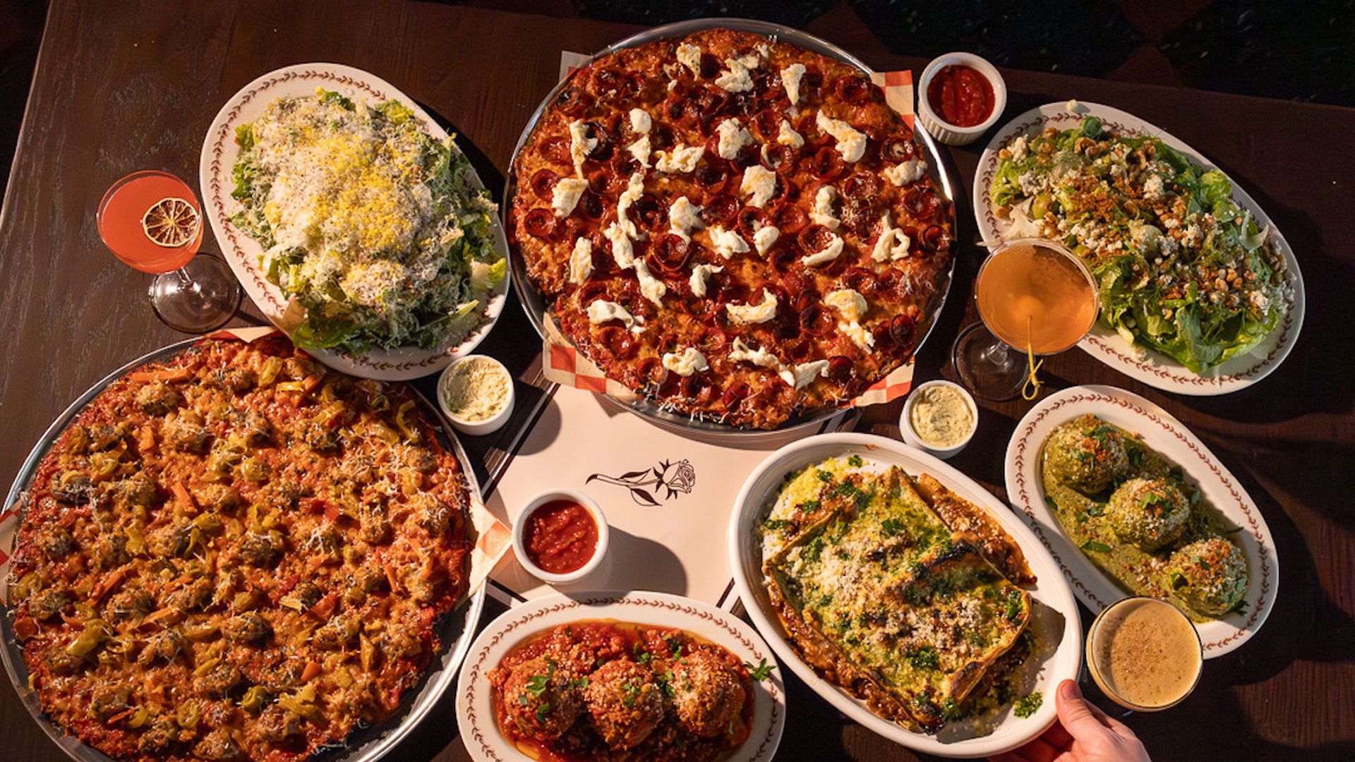 Top-down view of a table with multiple dishes: pepperoni pizza with ricotta, a large meat-and-vegetable pizza, two salads, meatballs in sauce, a baked casserole, and two colorful cocktails.
