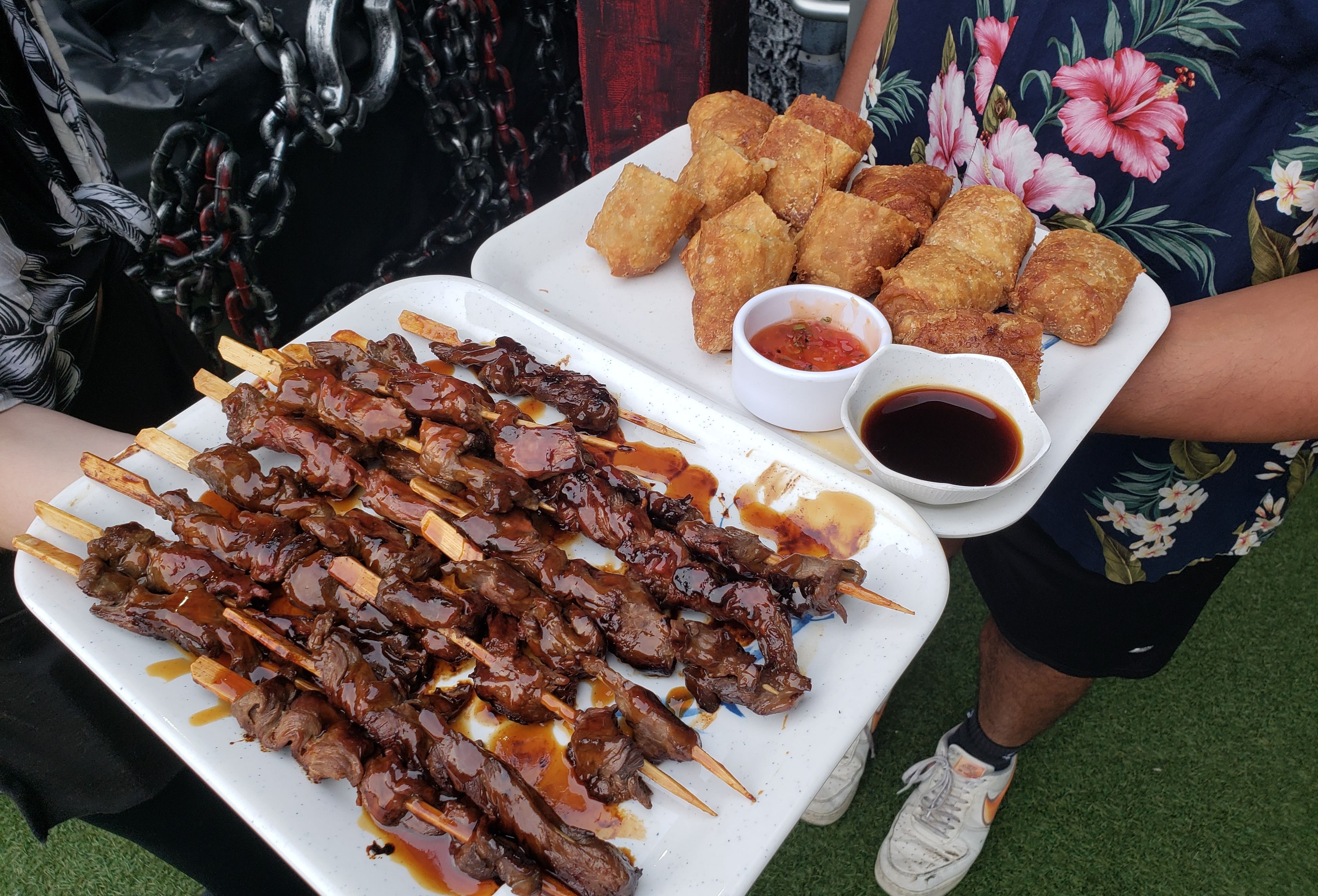 Meat skewers on white plate next to egg rolls with two dishes of sauce on white plate.