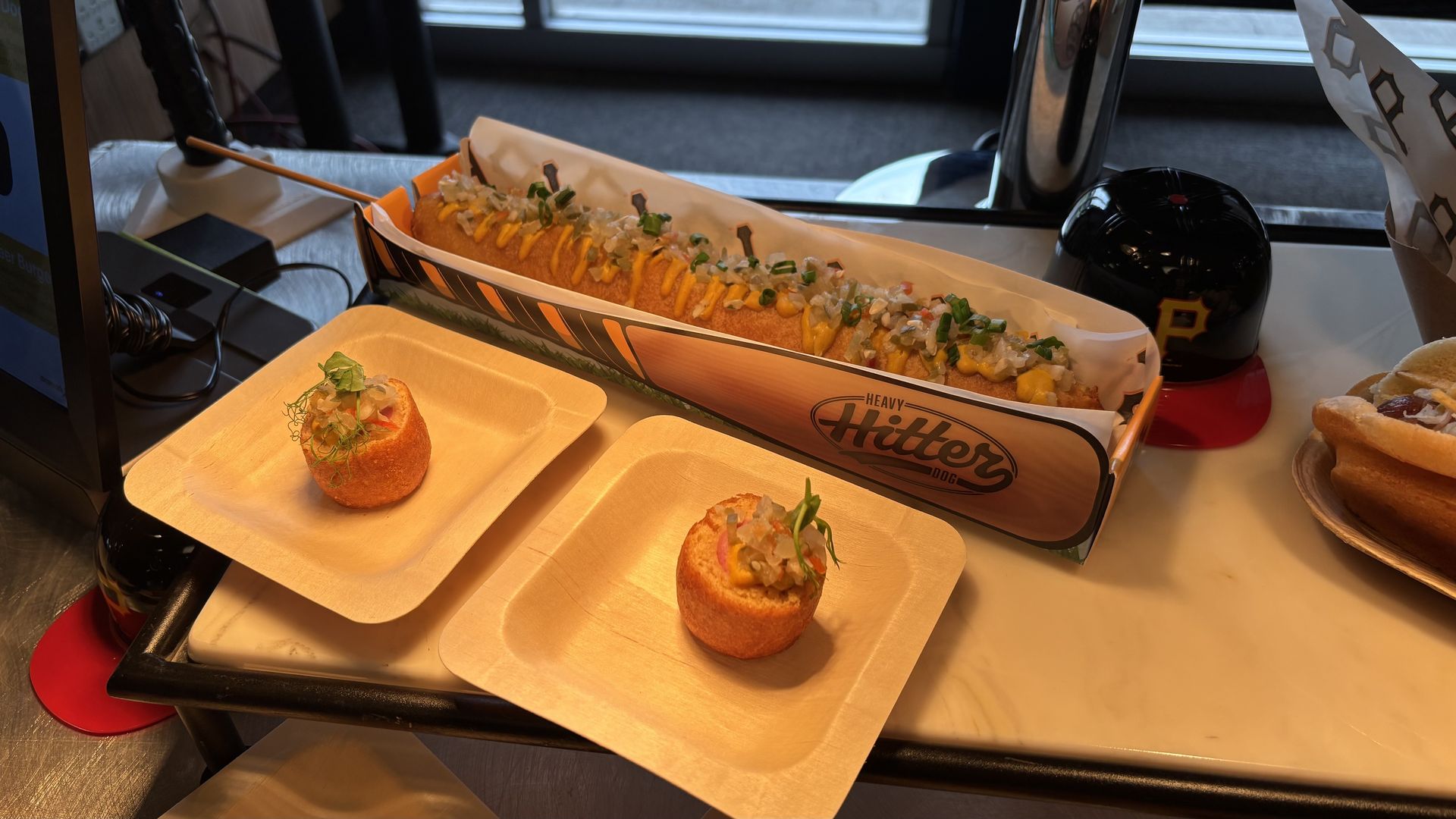 Long loaded hot dog in a paper tray topped with diced onions and herbs; two round fried bites on beige plates nearby. A black cap with a yellow P rests nearby, with a pastry on the right.