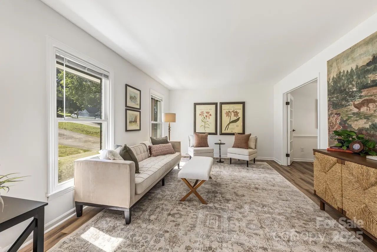 Bright living room with beige sofa, two armchairs with brown pillows, a patterned rug, large windows, framed botanical prints, and woven cabinet with plants on top.