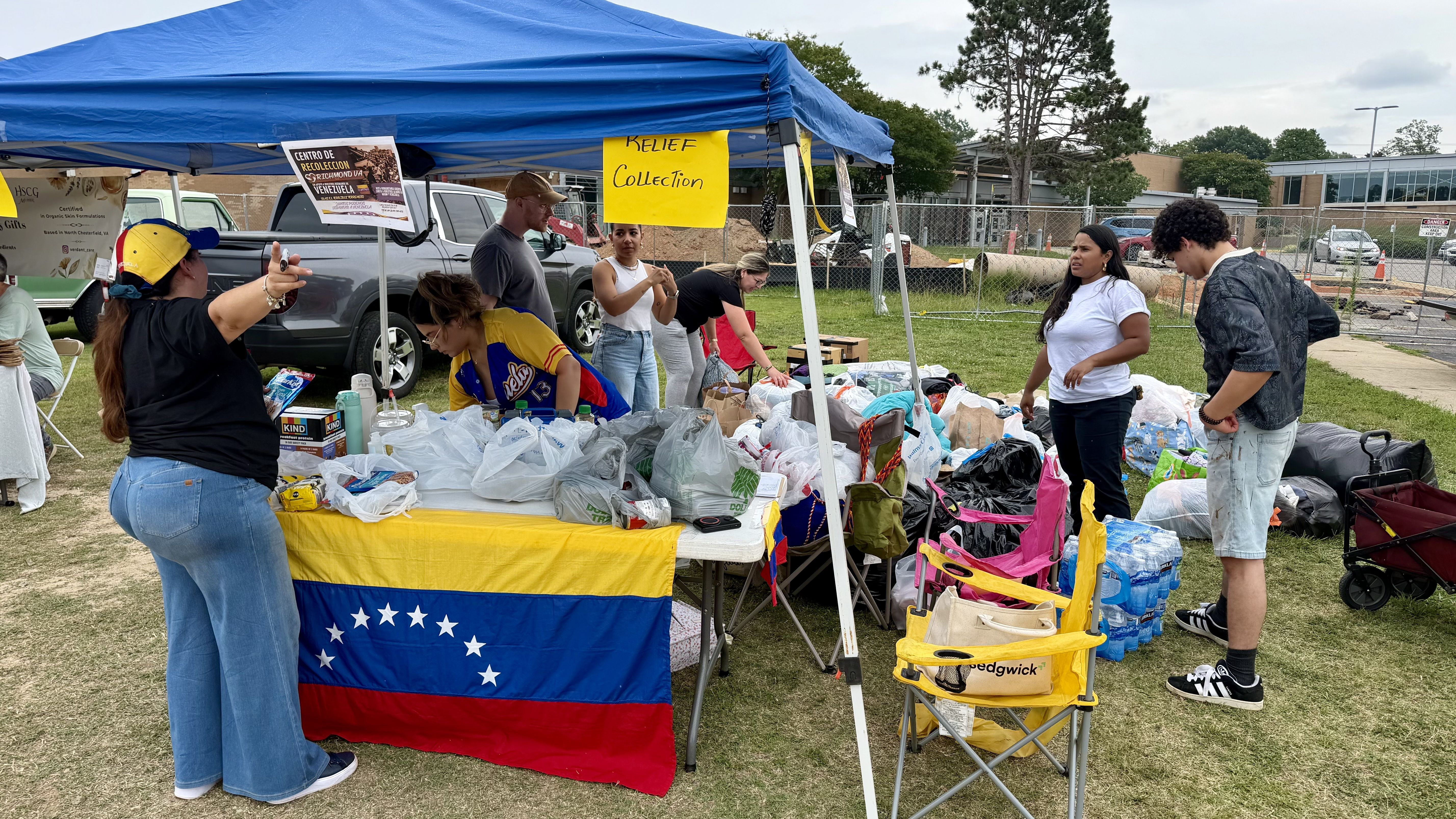 Outdoor relief drive under a blue canopy. Volunteers sort clothes and supplies on a table draped with a Venezuelan flag; a yellow sign reads "Relief Collection." A chain-link fence and campus buildings are in the background.