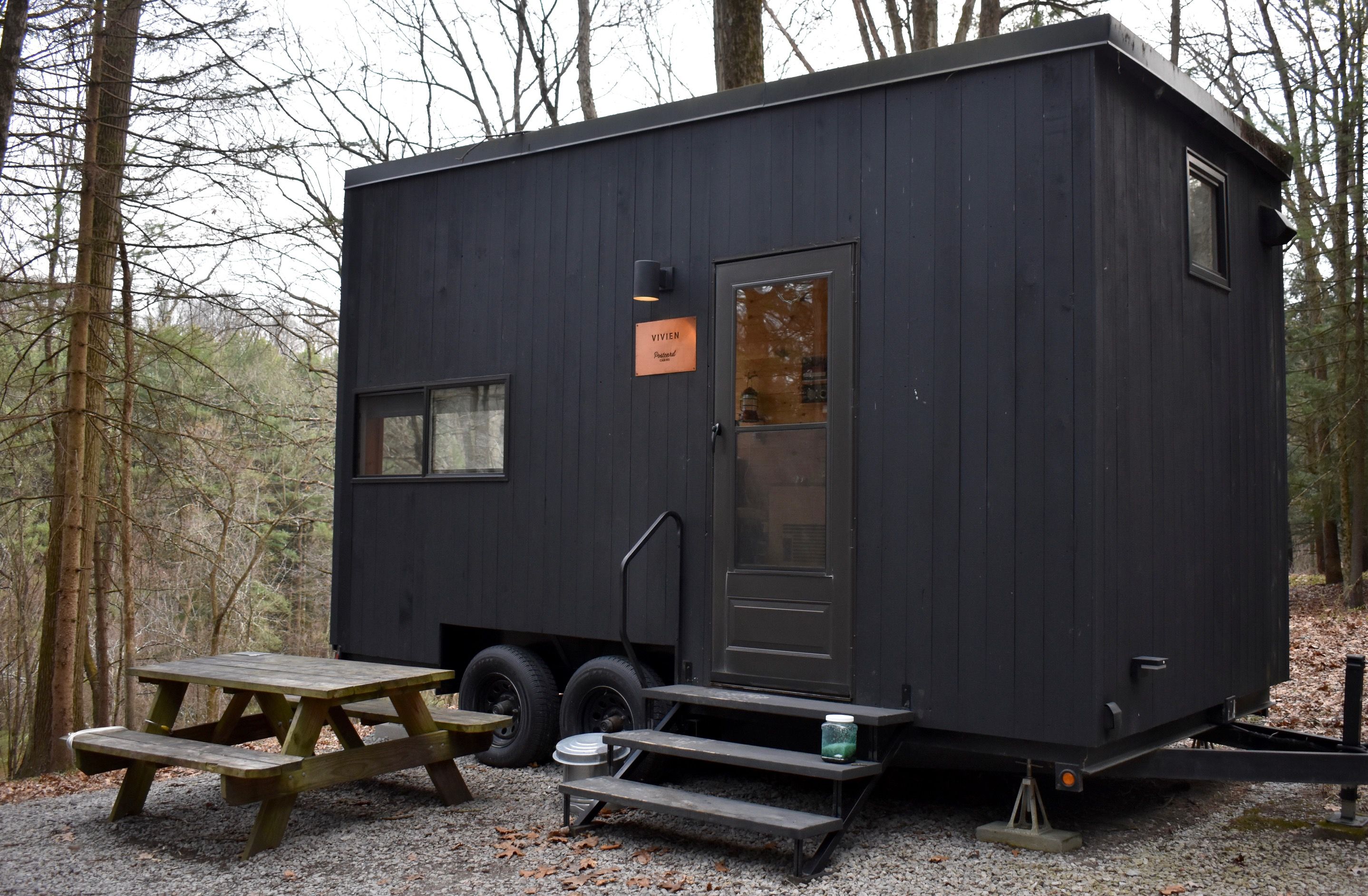 A dark blue tiny house on a trailer sits in a forest clearing. Gravel ground with a wooden picnic table outside. A door with steps and a small windowed wall, surrounded by bare trees.