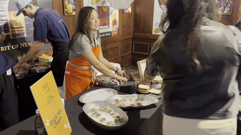 A woman moves two oysters from one platter to another as someone walks up to the table to take an oyster.