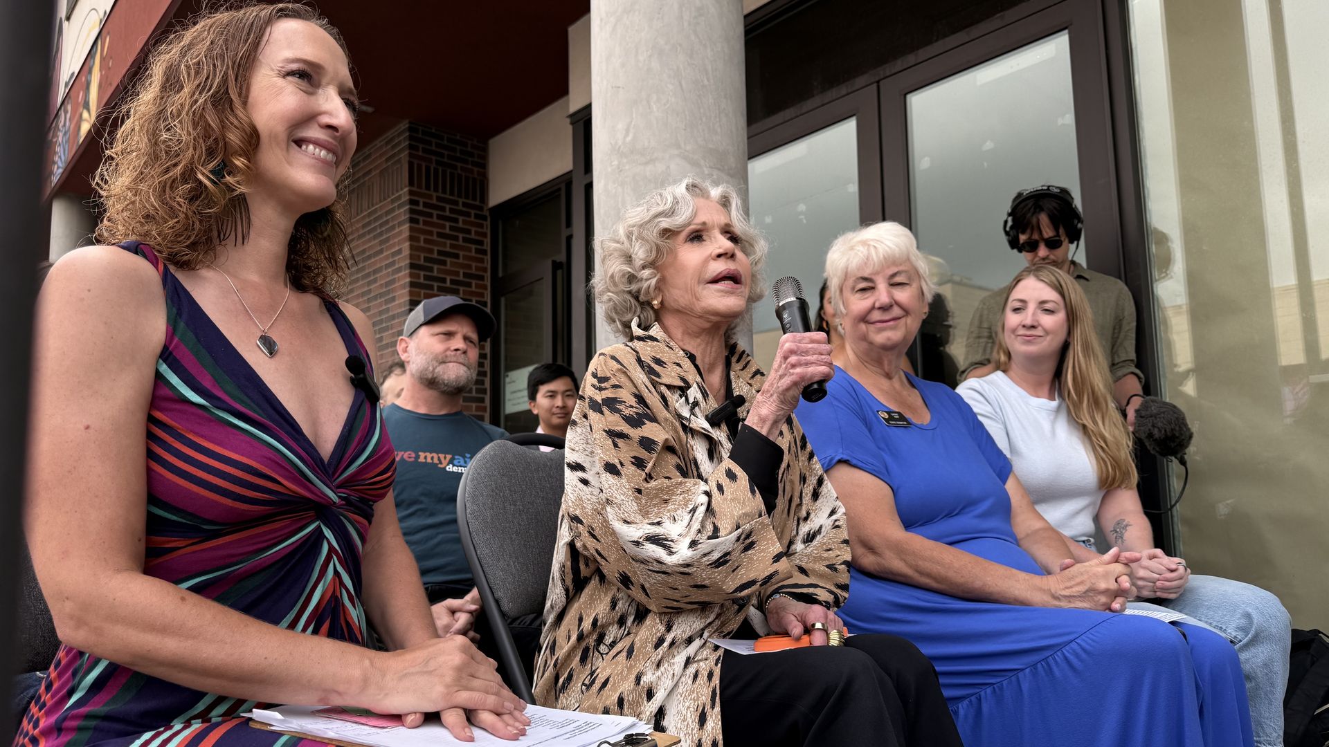 Group of people sitting outside a storefront; left foreground woman in a colorful striped dress smiles, center woman in leopard print jacket holds a microphone, others watch.