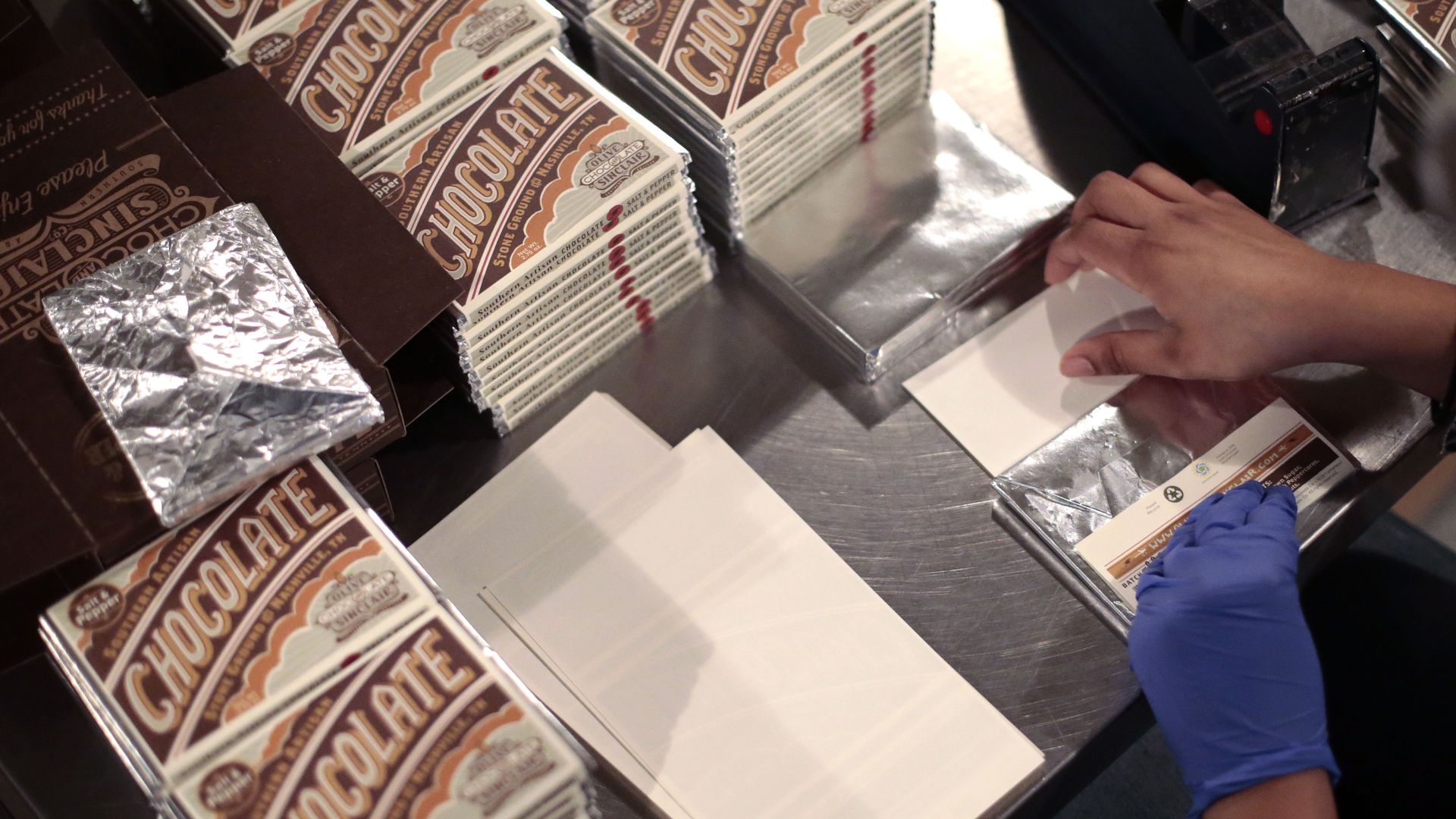 Hands shown wrapping chocolate bars in paper wrappers. Finished packaged bars are stacked around the table.