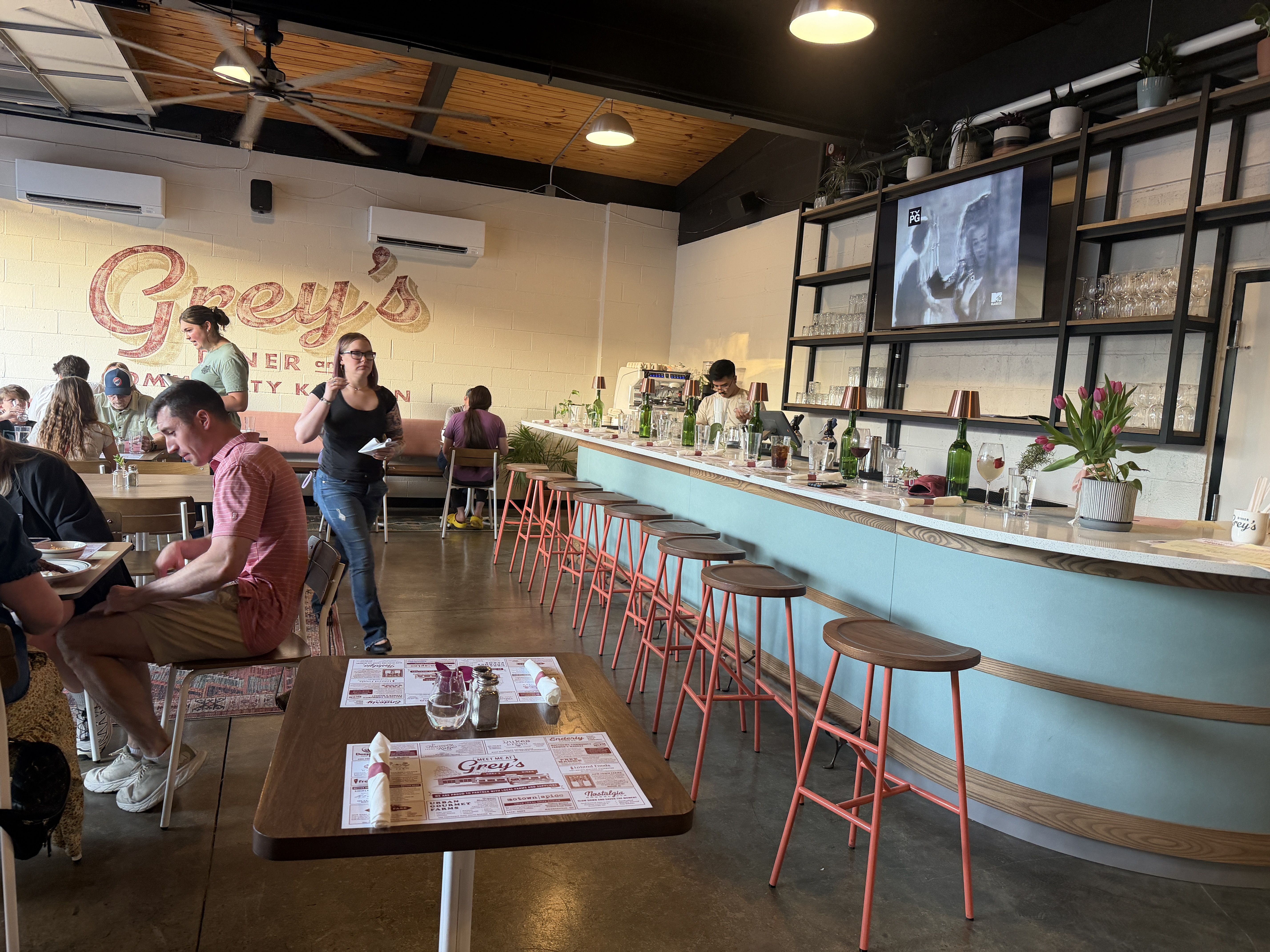 Casual cafe interior with a turquoise counter and pink stools along the right. Diners at tables on the left, white brick wall with a red cursive sign, ceiling fans, potted plants, and a mounted TV.