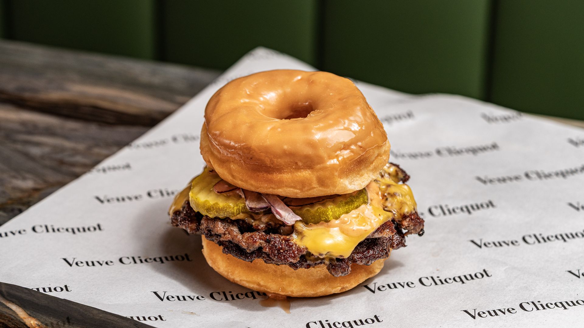 Close-up of a donut-bun burger: two beef patties with melted cheese, pickles, and bacon on parchment printed "Veuve Clicquot" beside a wooden table.
